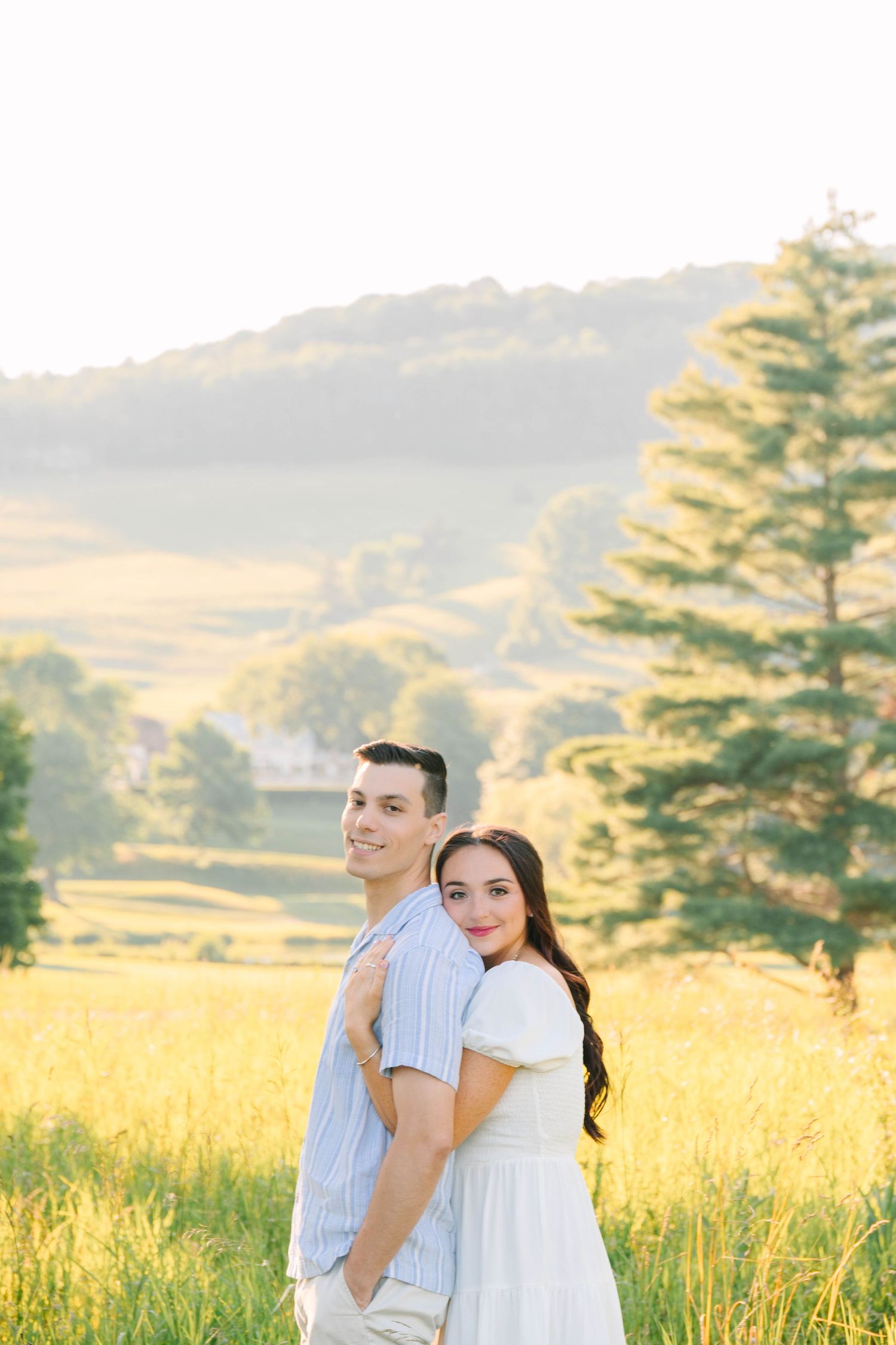 A couple poses back-to-back in a sunlit meadow with rolling hills and a pine tree in the background.