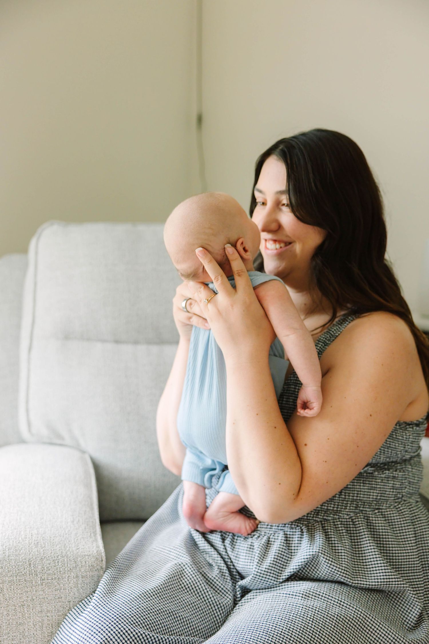 Smiling dark-haired mother holding newborn baby close to her face while sitting on a gray sofa.
