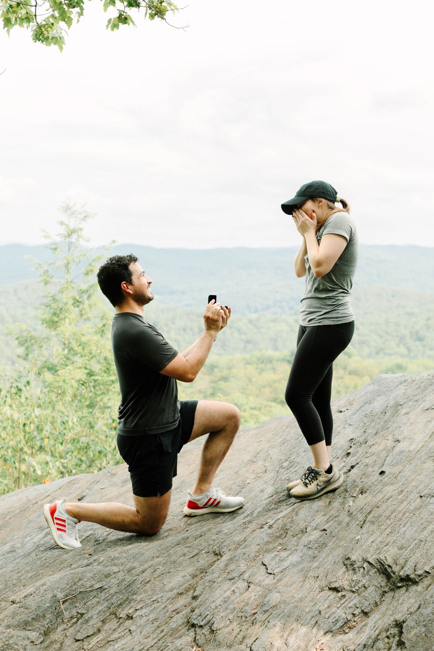 Man proposes on one knee holding ring box to surprised woman on rocky overlook with forest view.