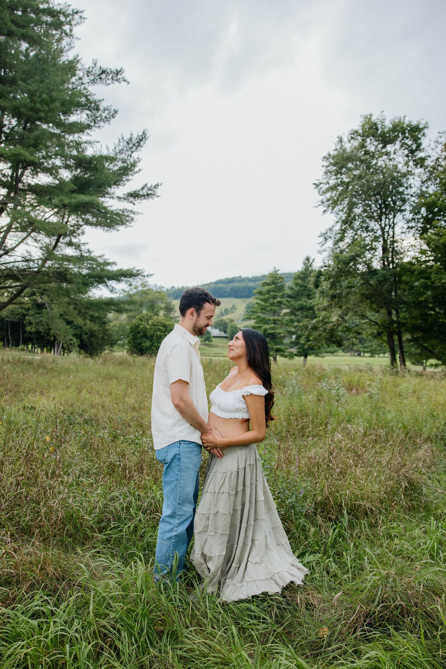 A couple stands face-to-face holding hands in a lush green meadow surrounded by trees on a cloudy day.