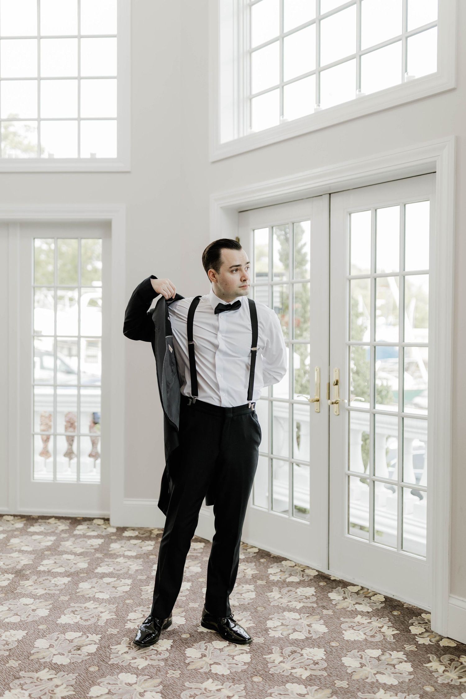 Groom in white shirt, suspenders and bow tie puts on black tuxedo jacket in bright windowed room.