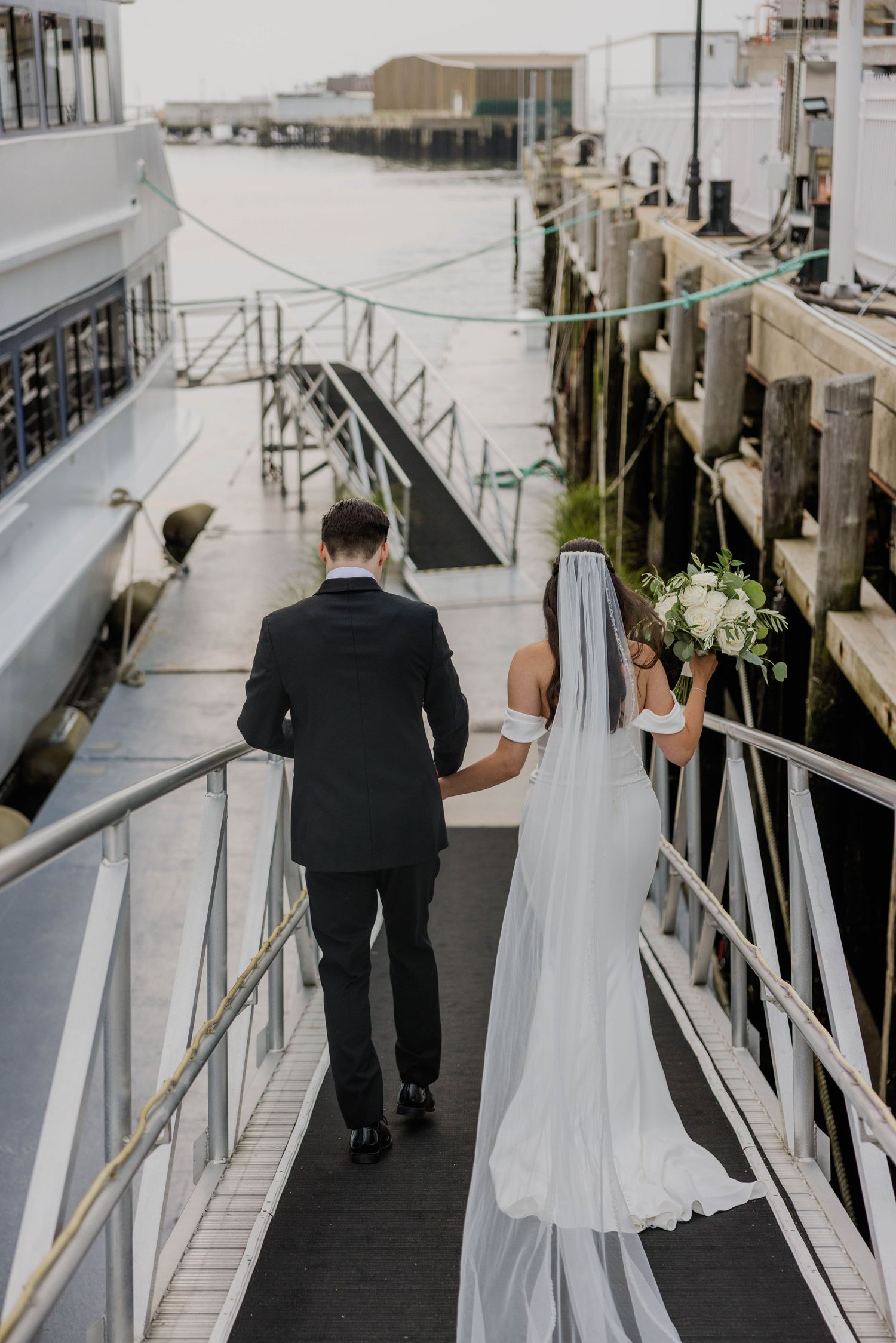 Bride and groom walk hand-in-hand down a waterfront dock, bride holding white floral bouquet in wedding dress and veil.