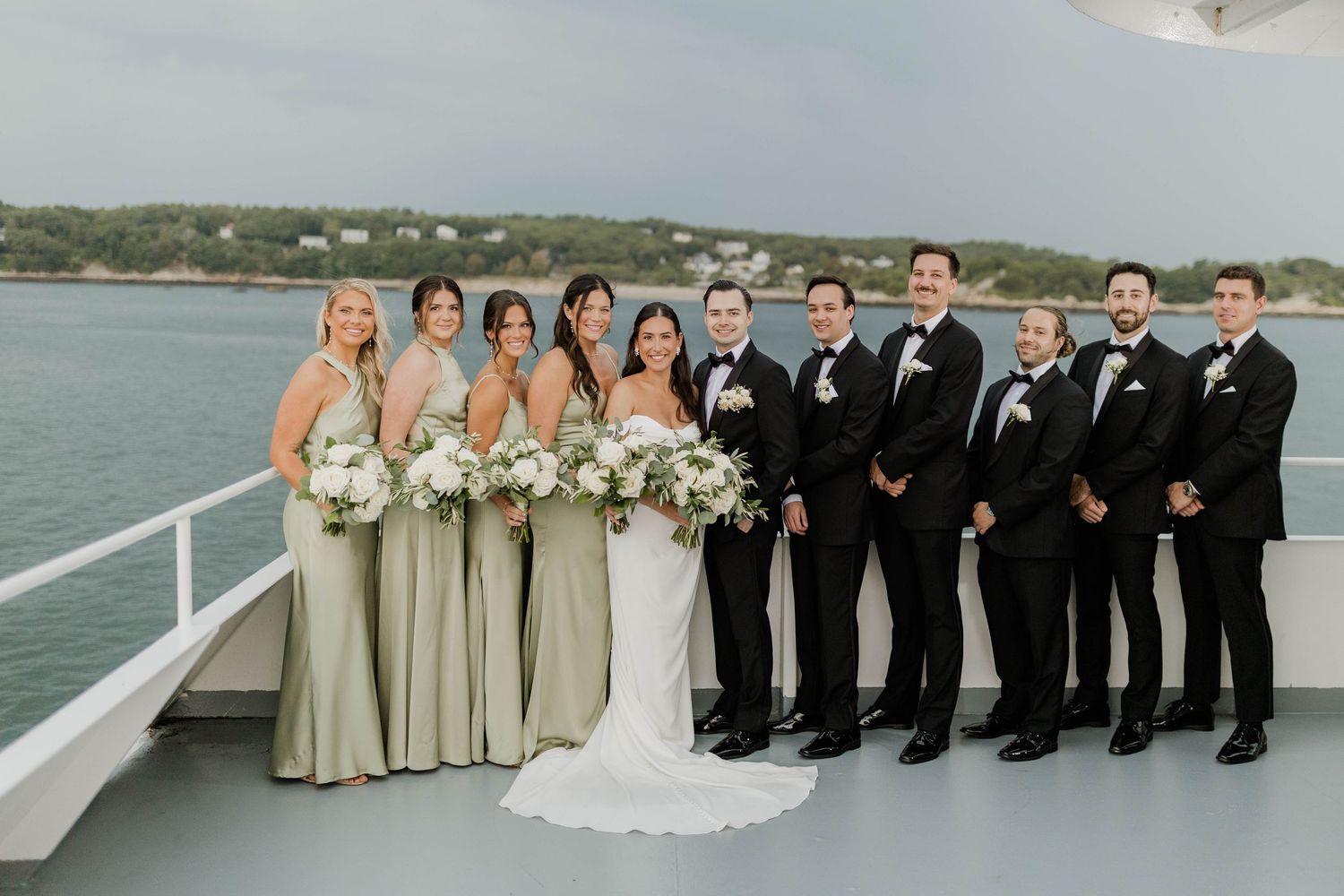 Wedding party posing on yacht deck with bridesmaids in sage green gowns and groomsmen in tuxedos.