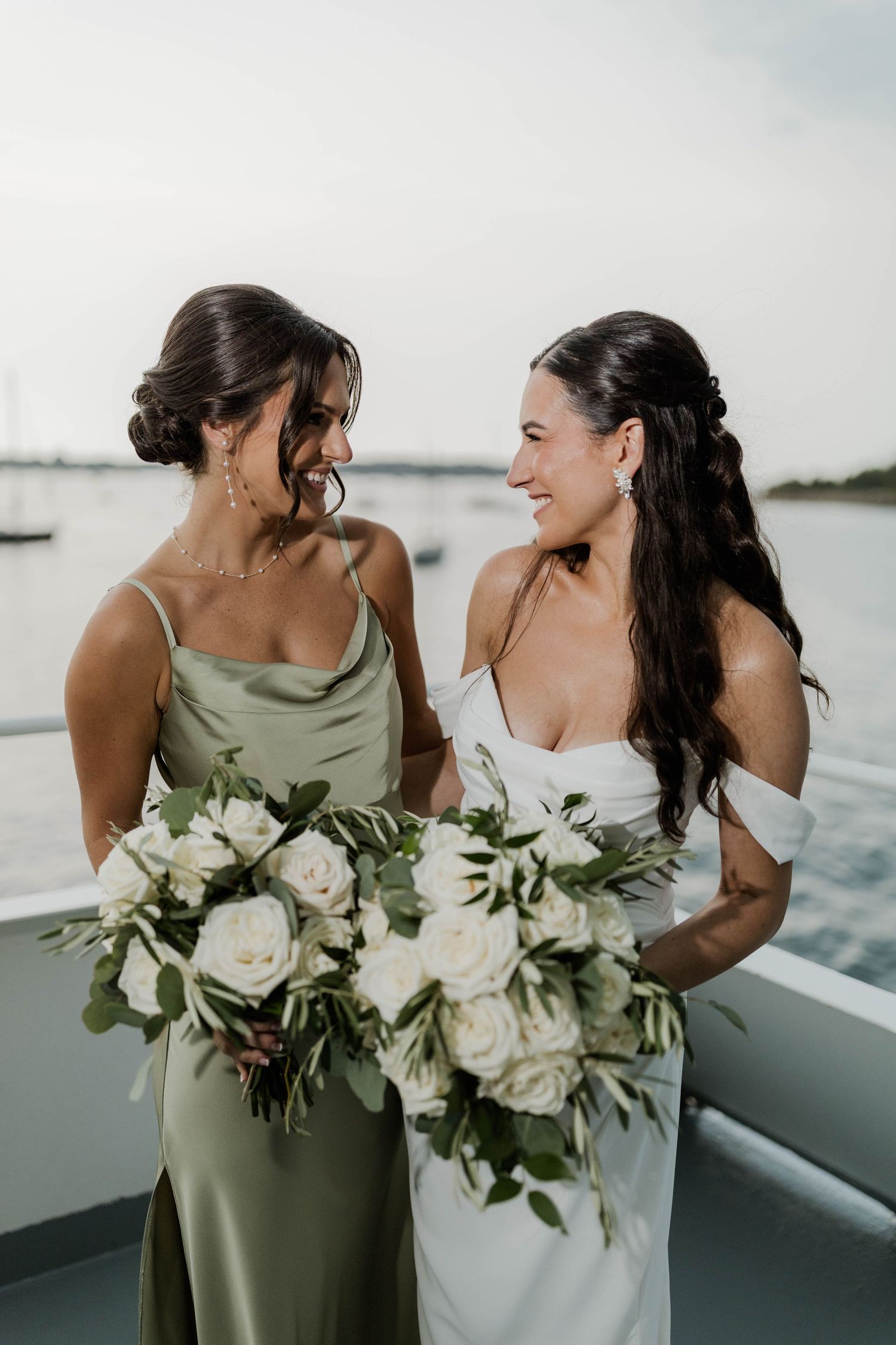 Bride and bridesmaid smile at each other on a boat, holding elegant white rose bouquets with greenery.