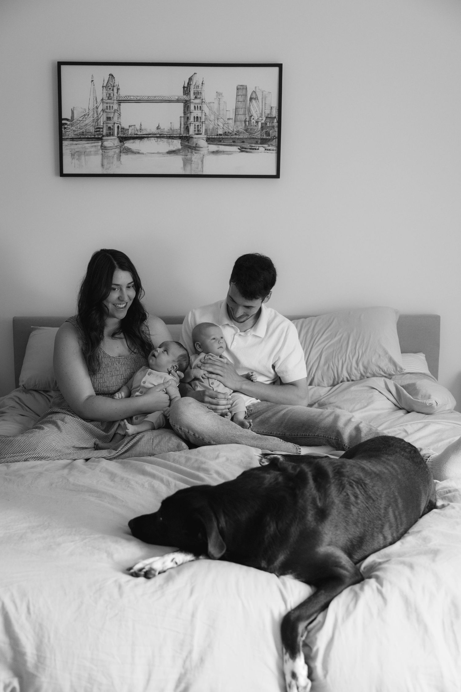 Black and white family photo of parents with twin babies and a dog relaxing on a bed together.