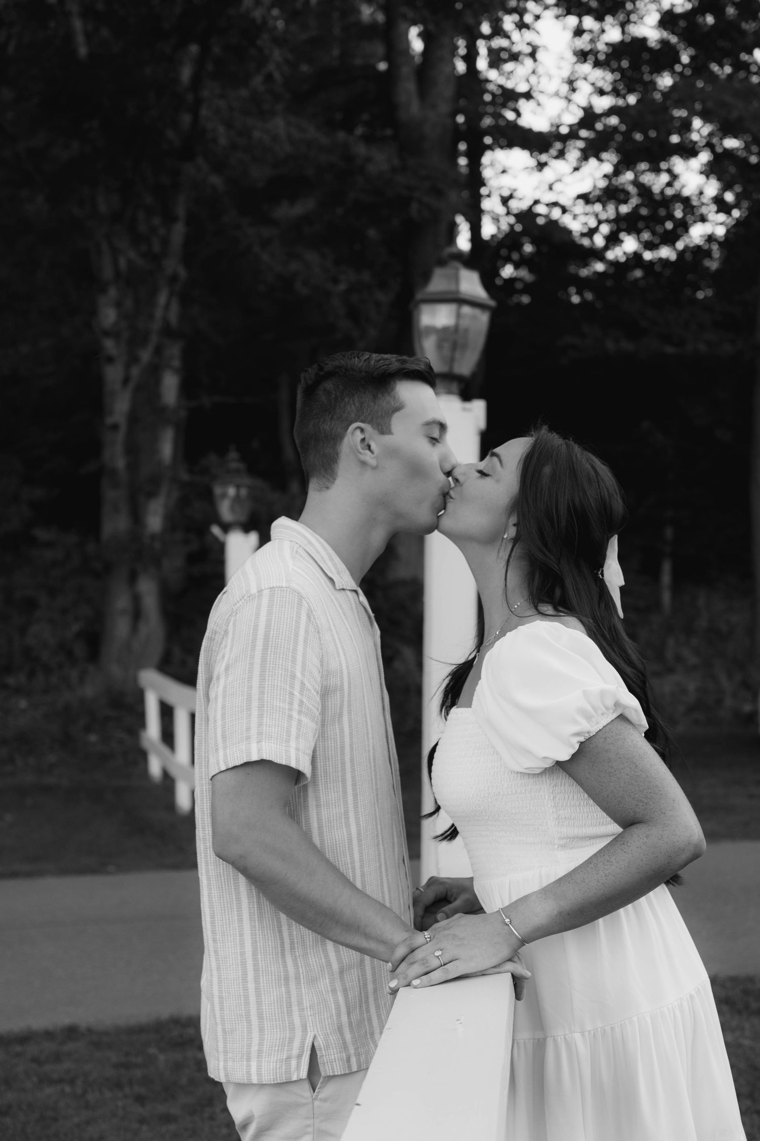 Black and white photo of a young couple sharing a romantic kiss outdoors near trees and a lamp post.
