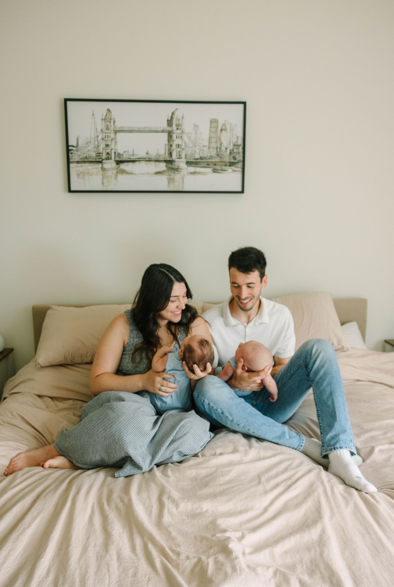 Happy young parents sitting on bed smiling at their newborn baby with London Bridge art above.