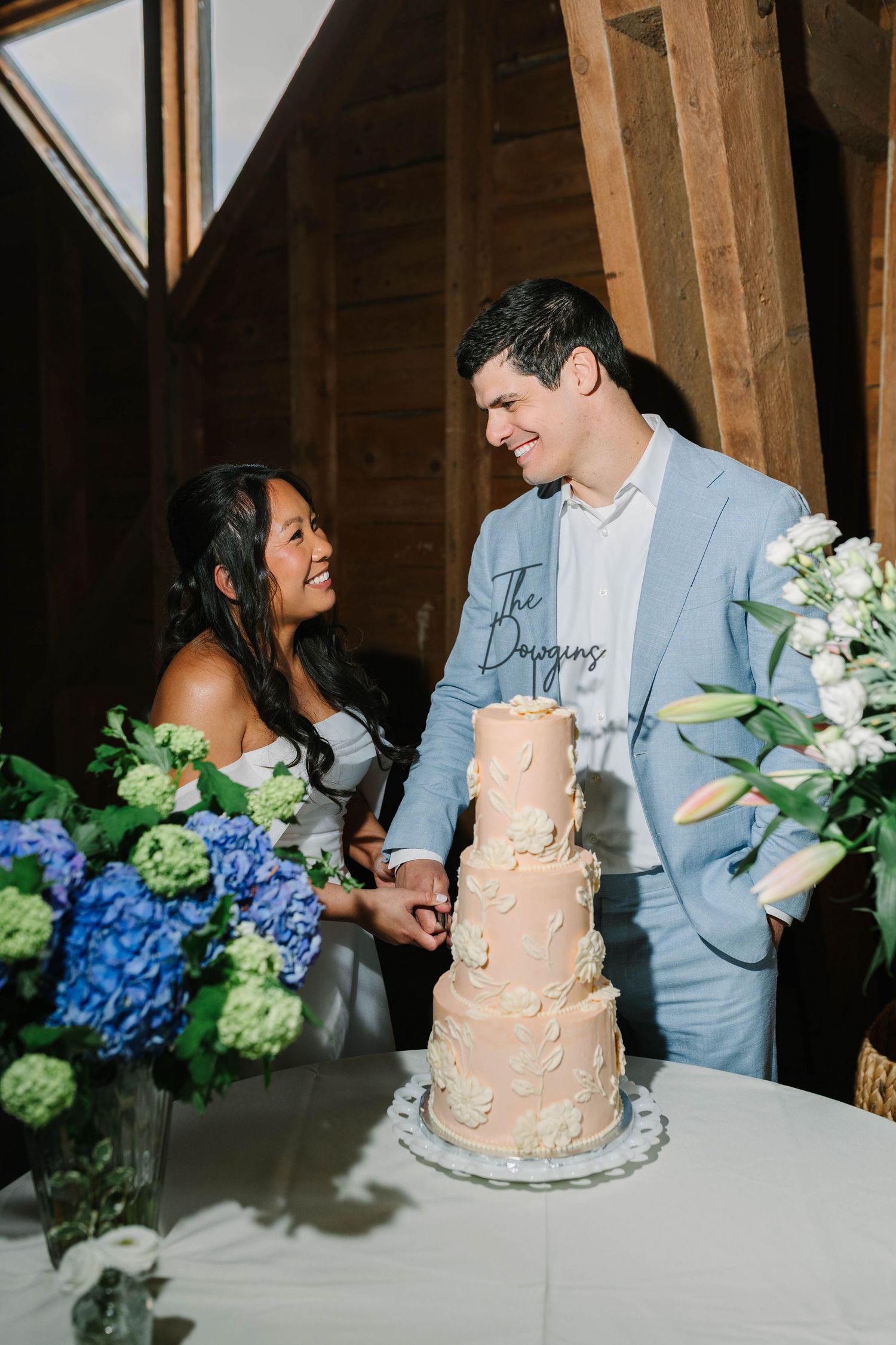 Bride and groom smile while cutting four-tier blush wedding cake surrounded by blue hydrangeas.