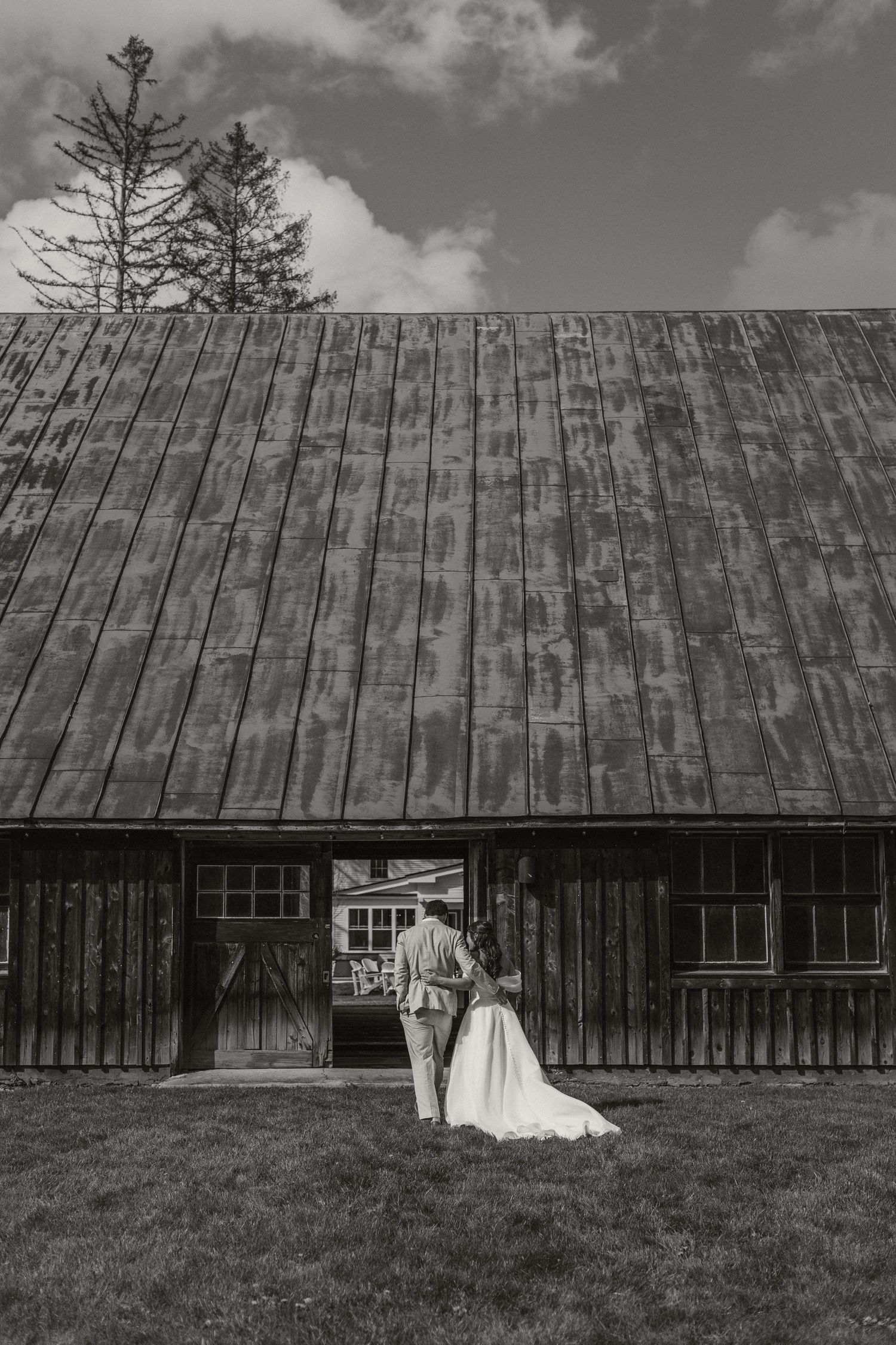 Bride and groom embrace at entrance of rustic wooden barn on green lawn in black and white photo.