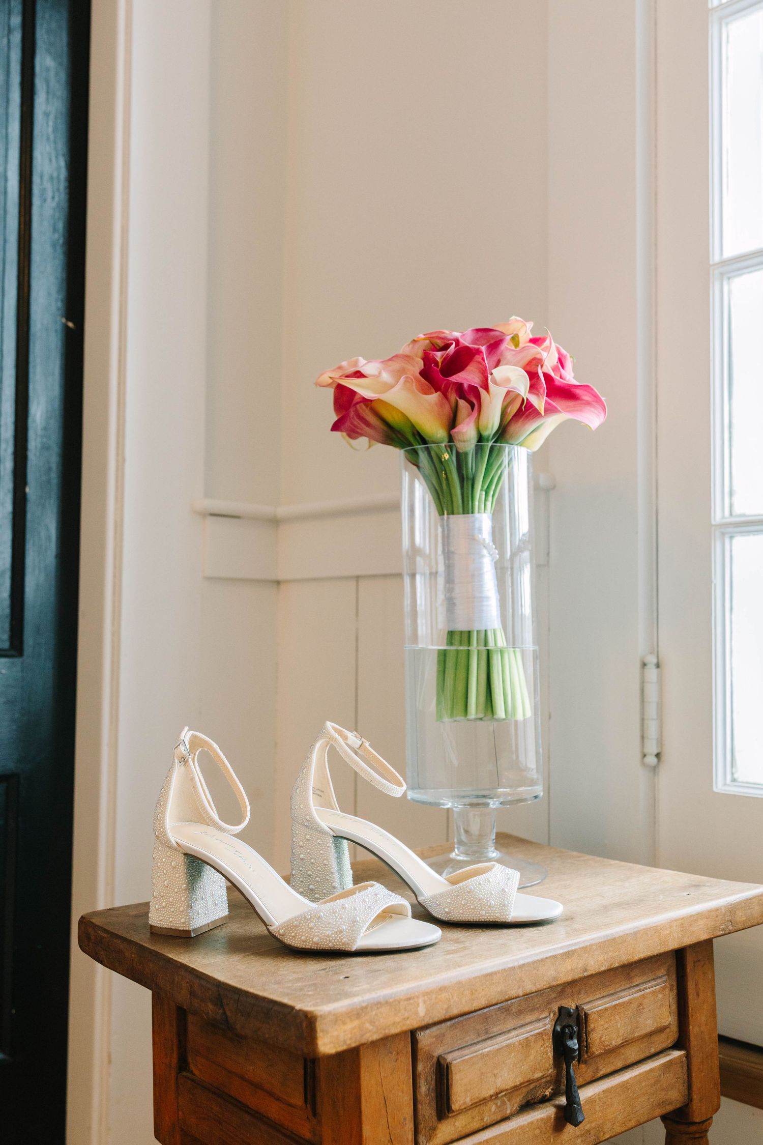 White bridal heels displayed on wooden table beside pink calla lily bouquet in glass vase near window.