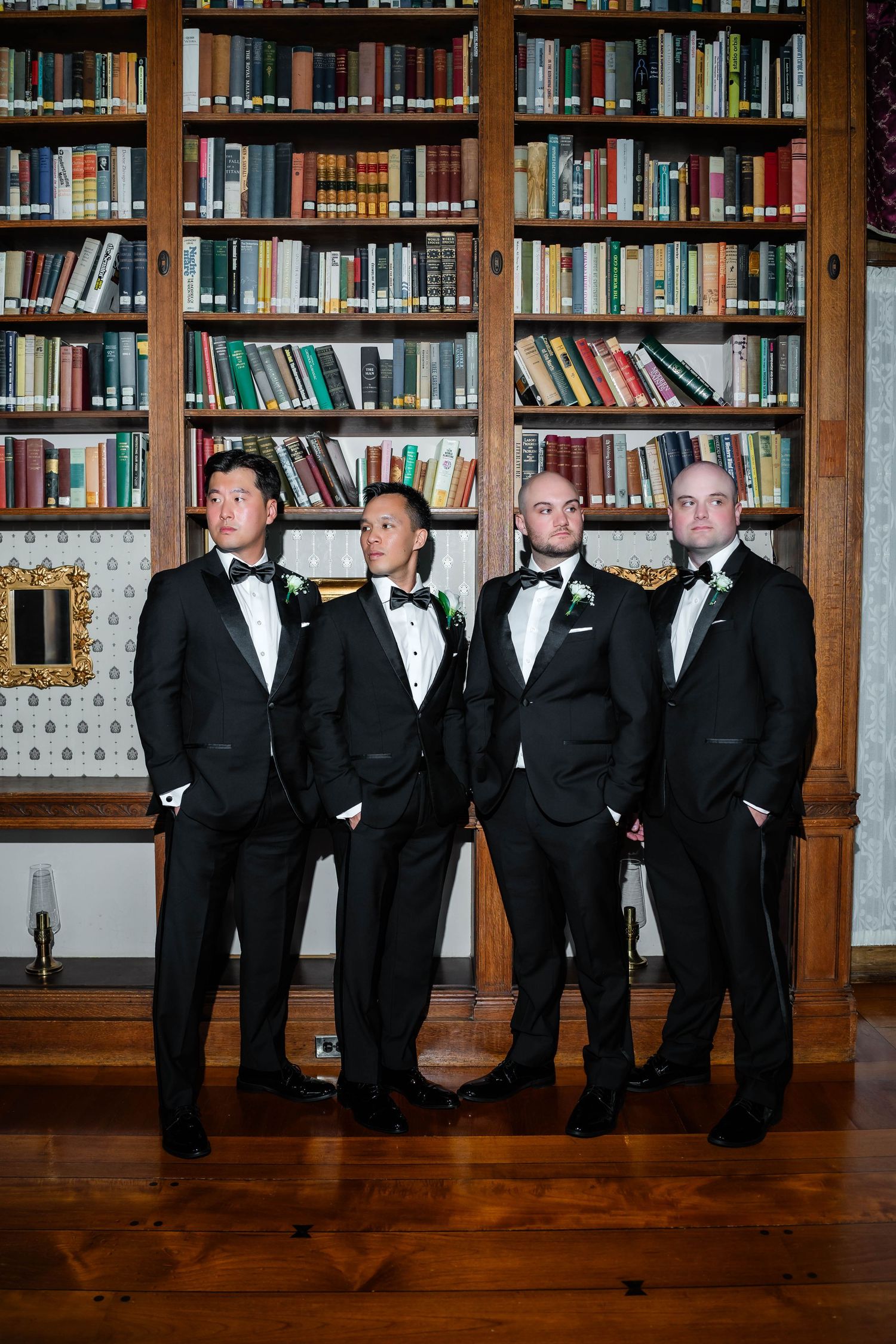 Four groomsmen in black tuxedos standing confidently before large wooden bookcase in library.