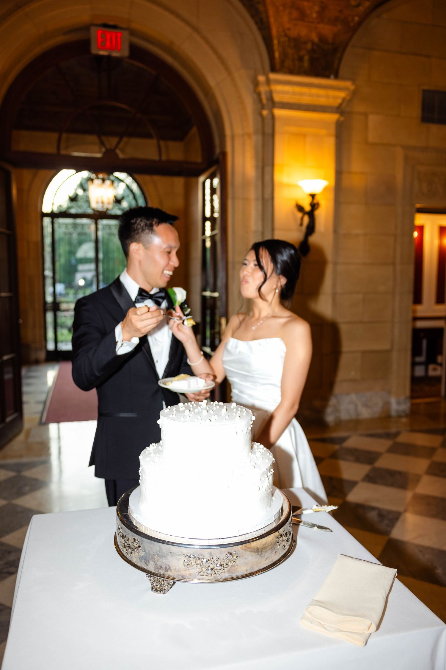 Smiling bride and groom cut elegant two-tier white wedding cake at reception.