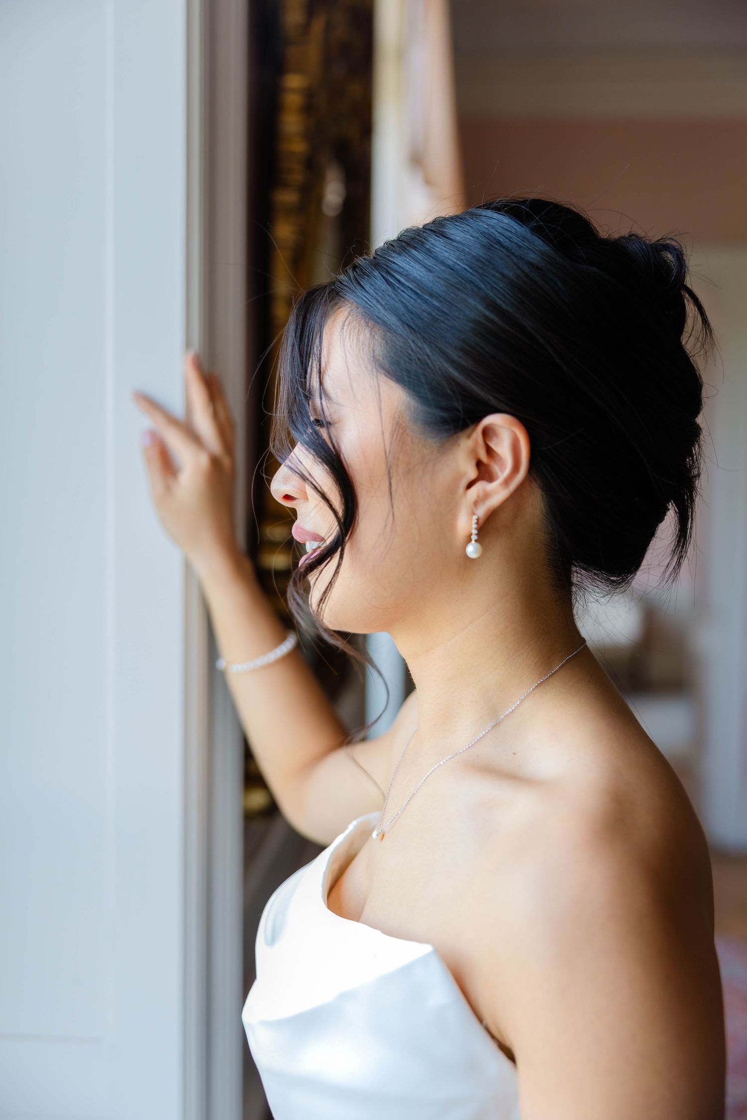 Bride in white strapless gown with pearl earrings and necklace gazing out a window on her wedding day.