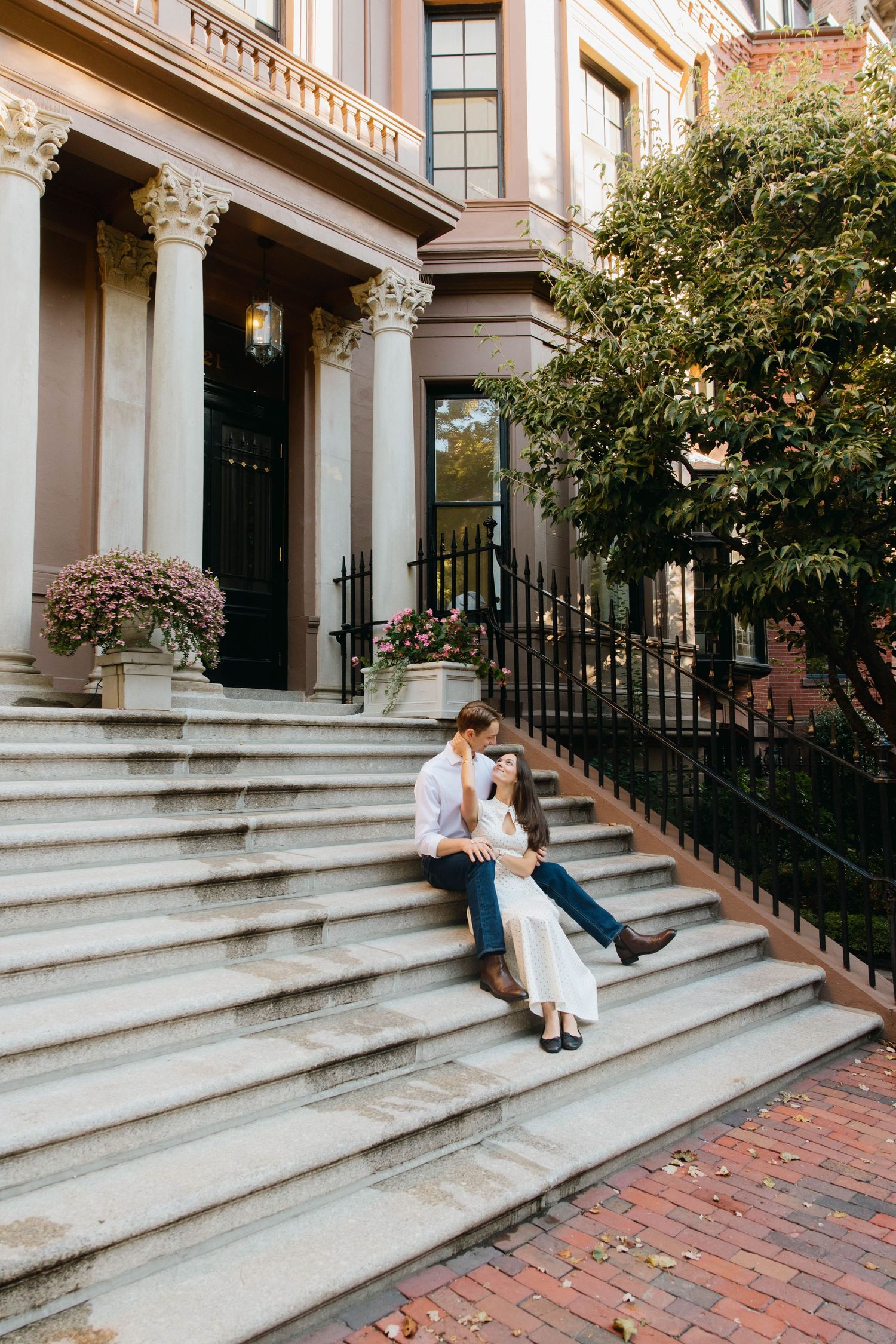 Couple sitting on brownstone steps in an urban neighborhood, surrounded by lush greenery and blooming flowers.