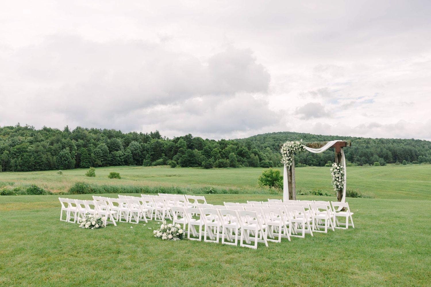 Elegant outdoor wedding ceremony setup with white chairs and a rustic floral arch on a green lawn.