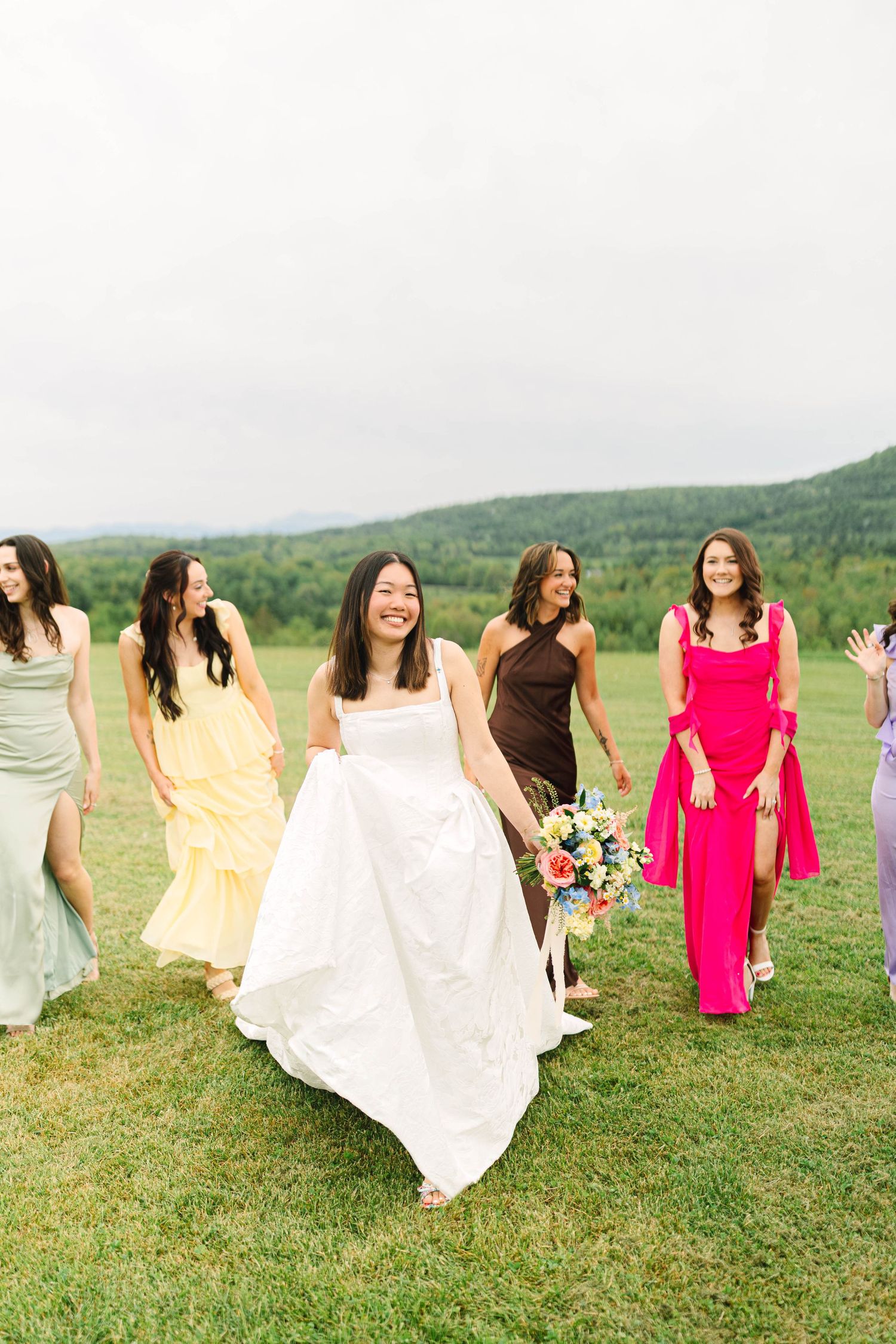 Joyful bride in white gown walks with bridesmaids in colorful mismatched dresses across a lush green mountain landscape.