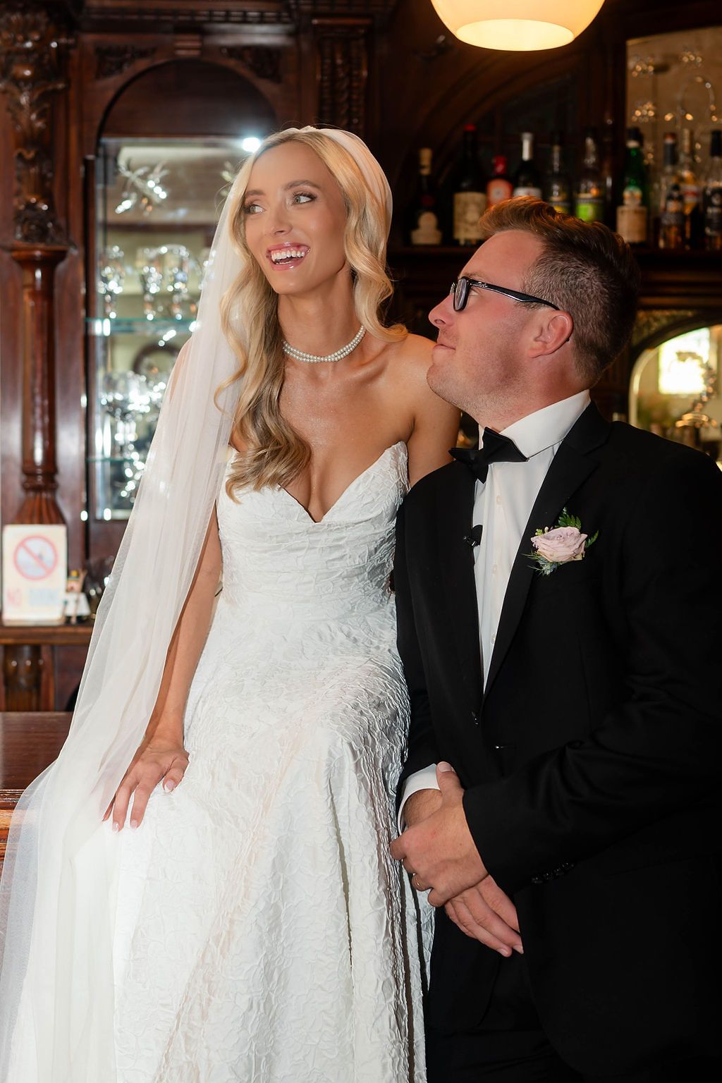 Smiling bride in lace gown and veil standing with groom in tuxedo at bar.