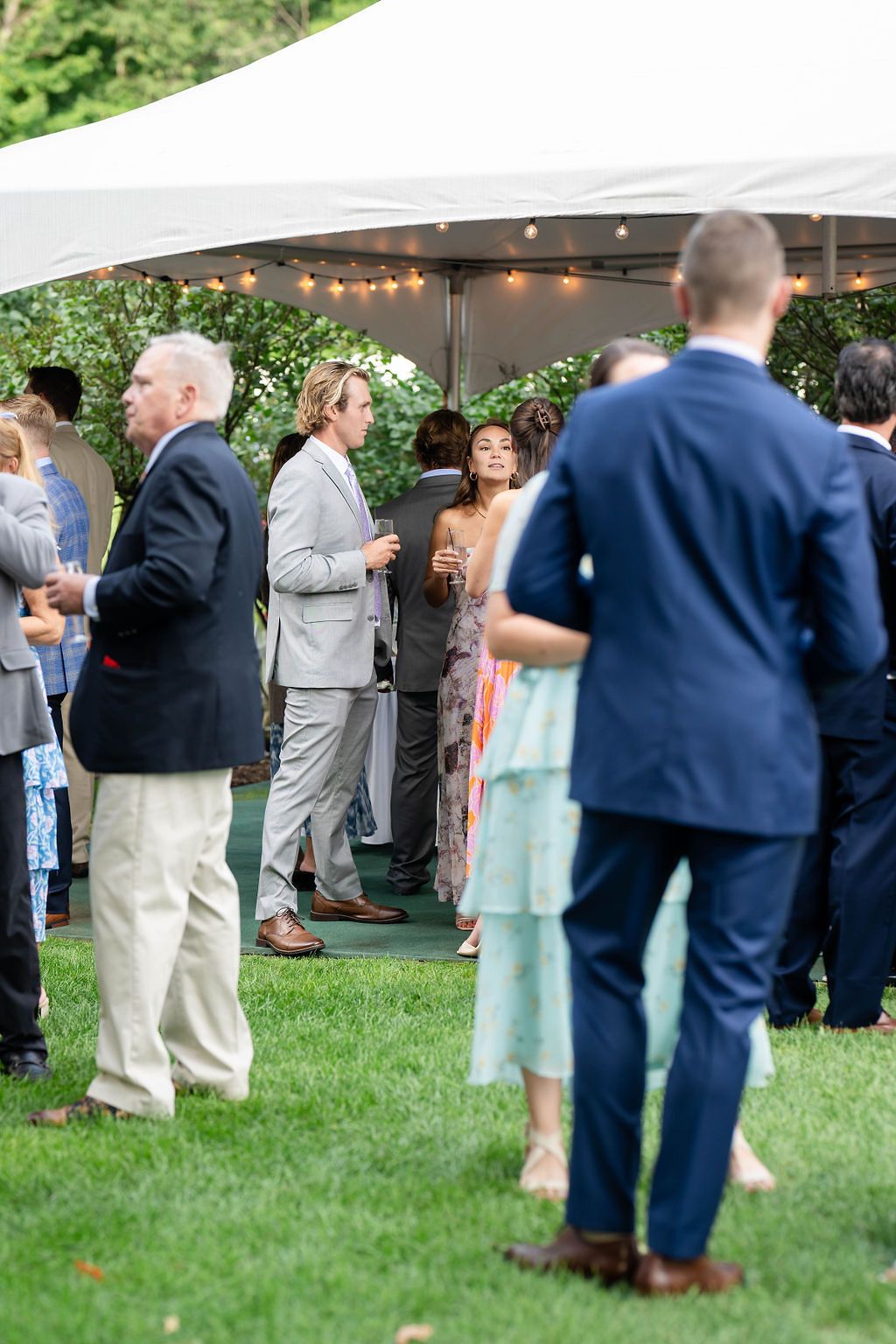 Wedding guests mingling on grass under a white tent with string lights at an elegant outdoor reception.