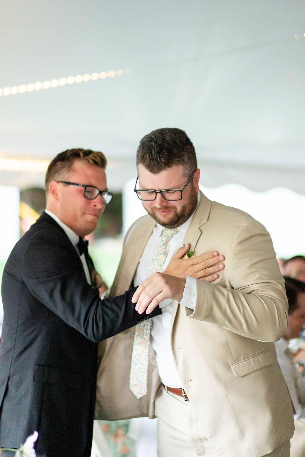 Groom in black suit pinning boutonniere on groomsman in tan suit at wedding reception.