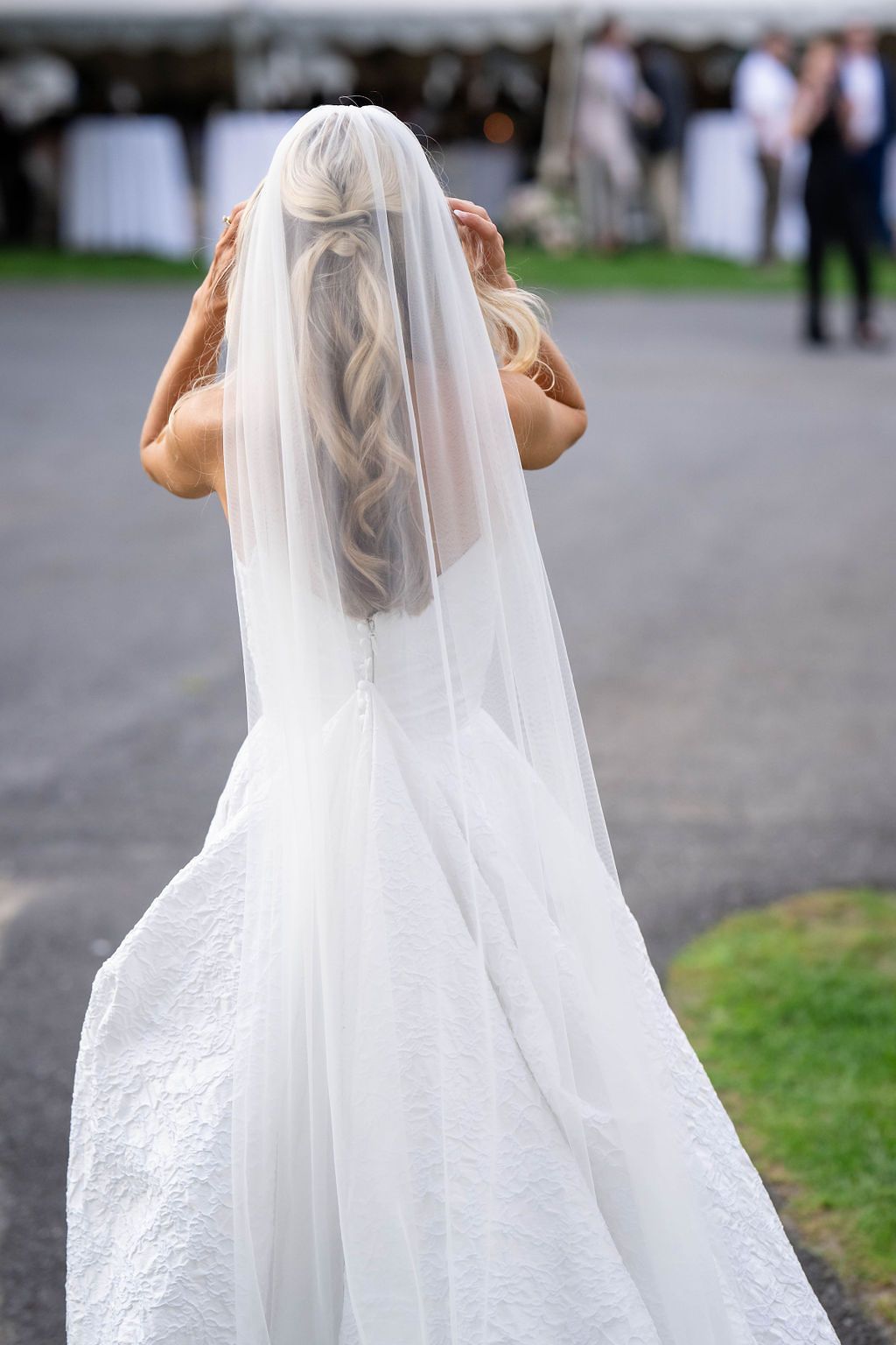 Rear view of bride in white gown and veil adjusting her braided hair at outdoor wedding venue.