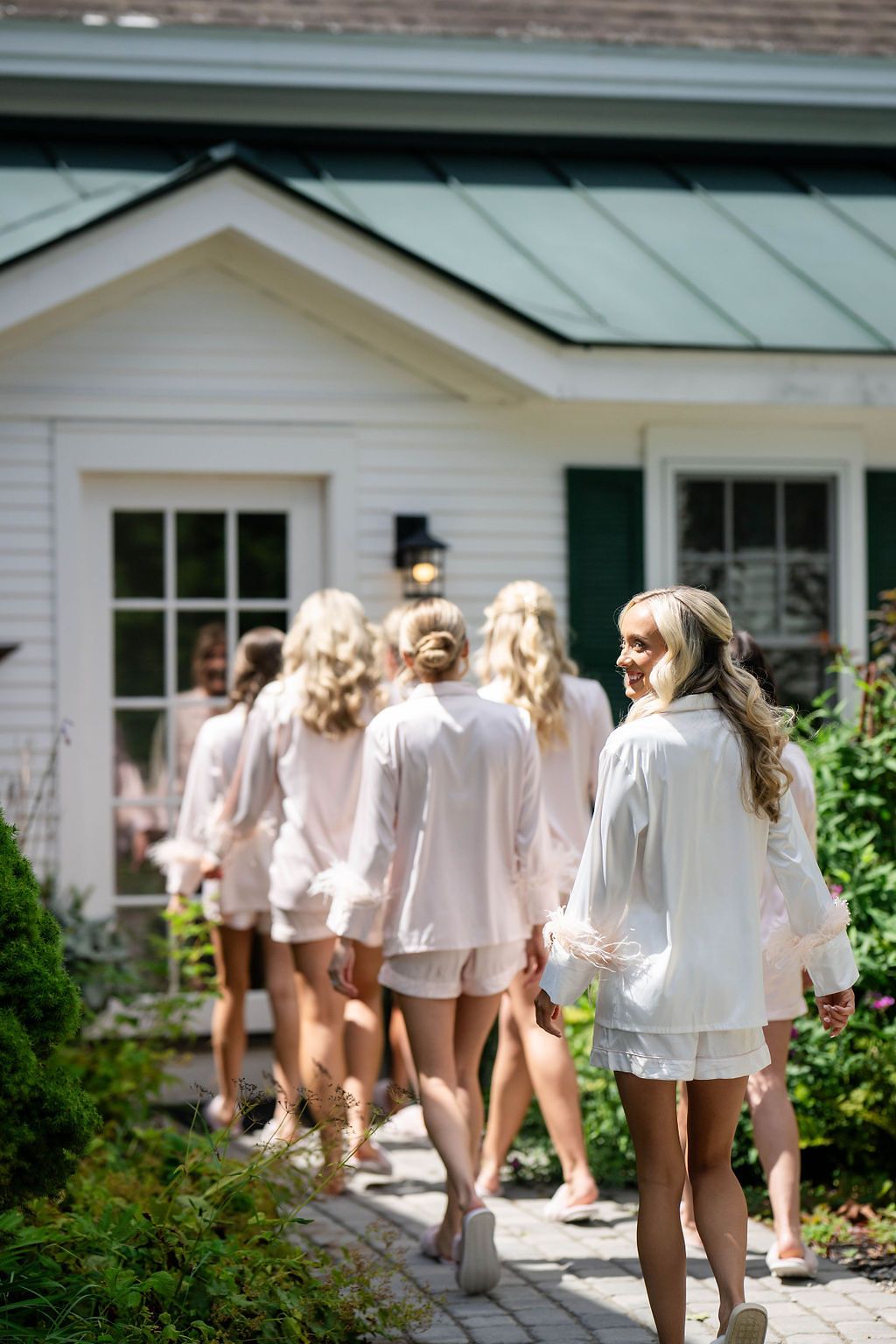 Bridesmaids in matching white satin pajama sets walking toward a white cottage during bridal party preparations.