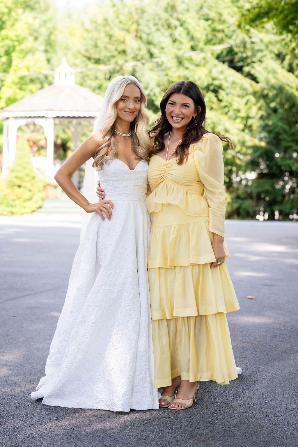Bride in white gown poses with guest in yellow tiered dress at outdoor wedding venue with gazebo.