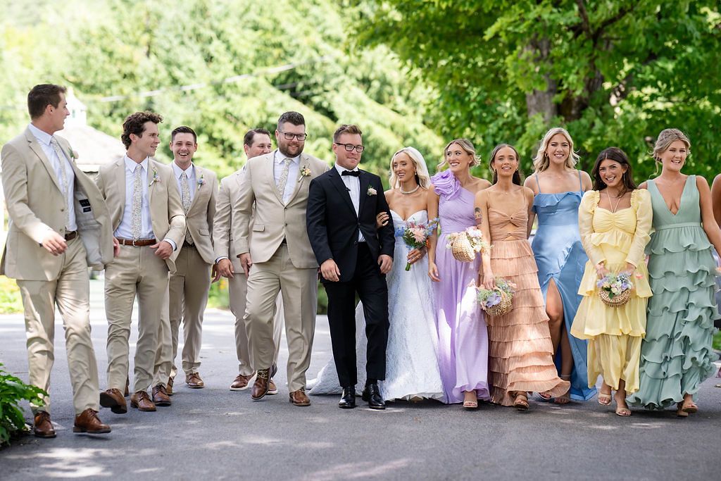 Wedding party walks together outdoors, groomsmen in tan suits and bridesmaids in colorful pastel dresses.