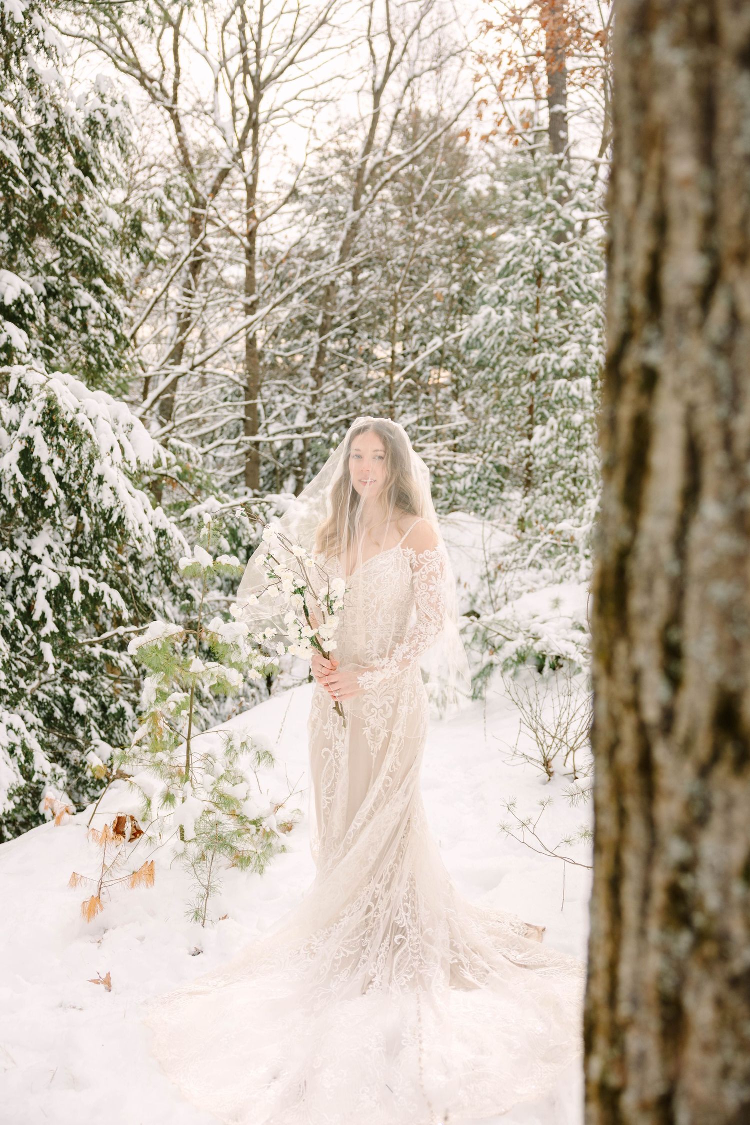Bride in lace gown and flowing veil stands holding white floral stems in a sunlit snow-covered forest.