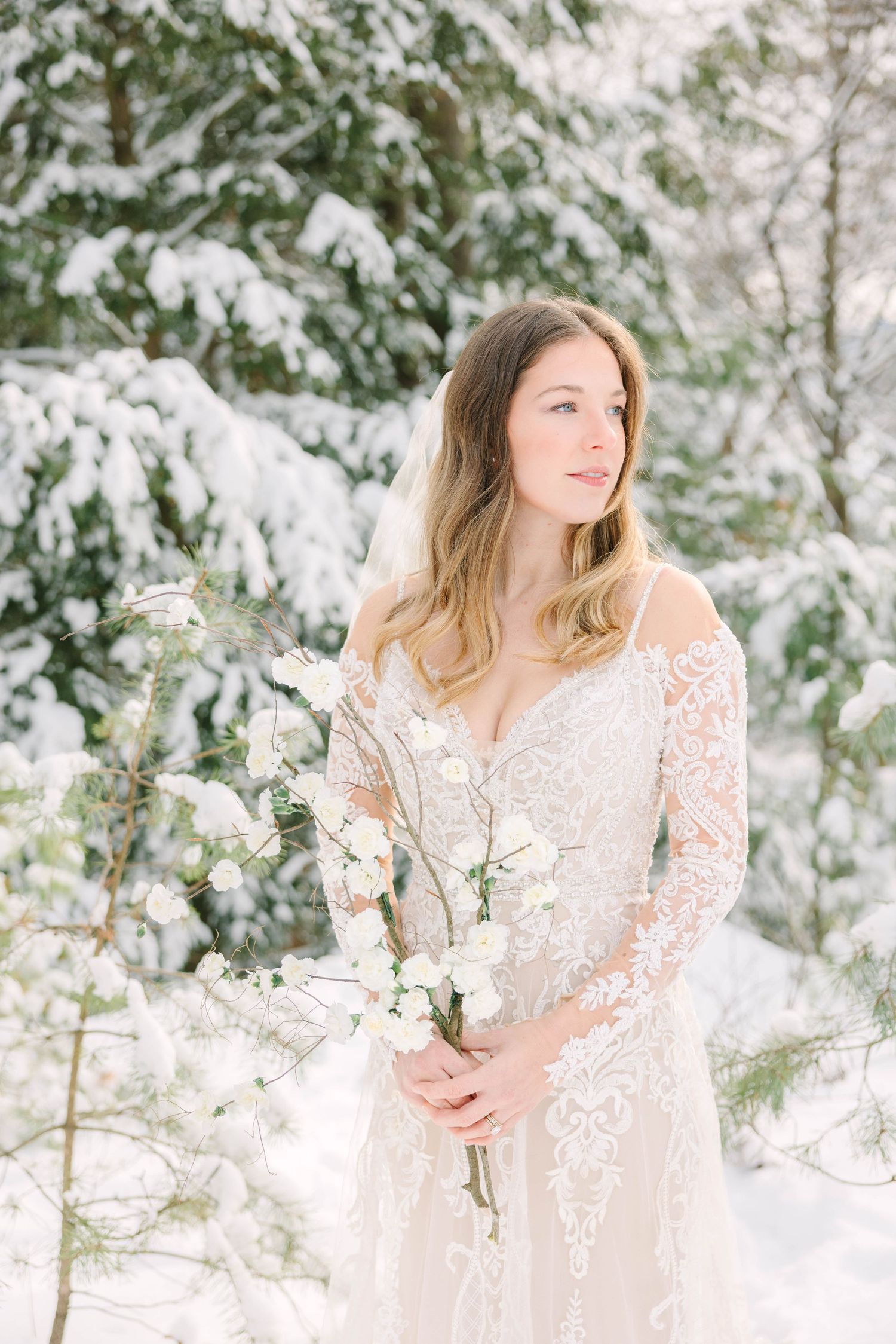 Bride in lace off-shoulder gown holding white floral bouquet in snowy winter forest setting.
