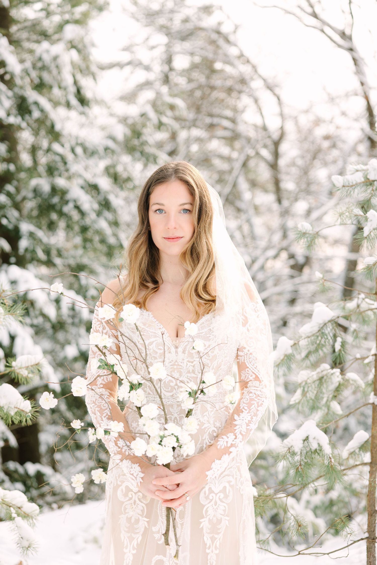 Bride in lace gown and veil holds white floral bouquet among snow-covered pine trees in winter forest.