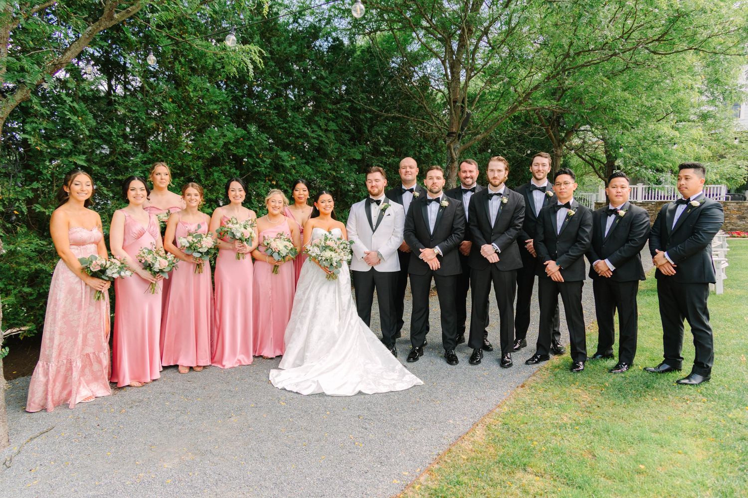 Full wedding party poses outdoors with bridesmaids in pink and groomsmen in black tuxedos.