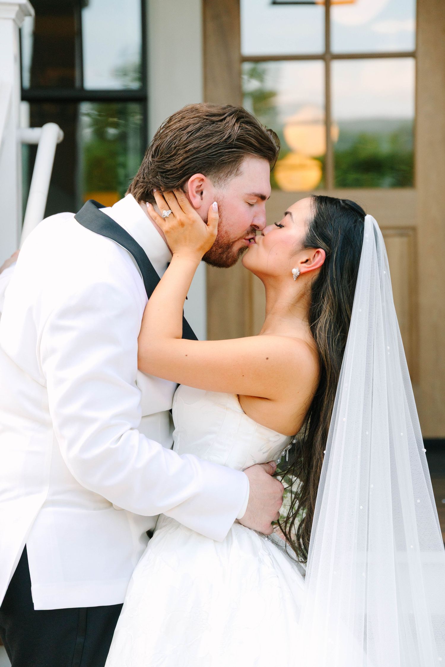 Bride in strapless gown and veil sharing a romantic kiss with groom in white tuxedo jacket.