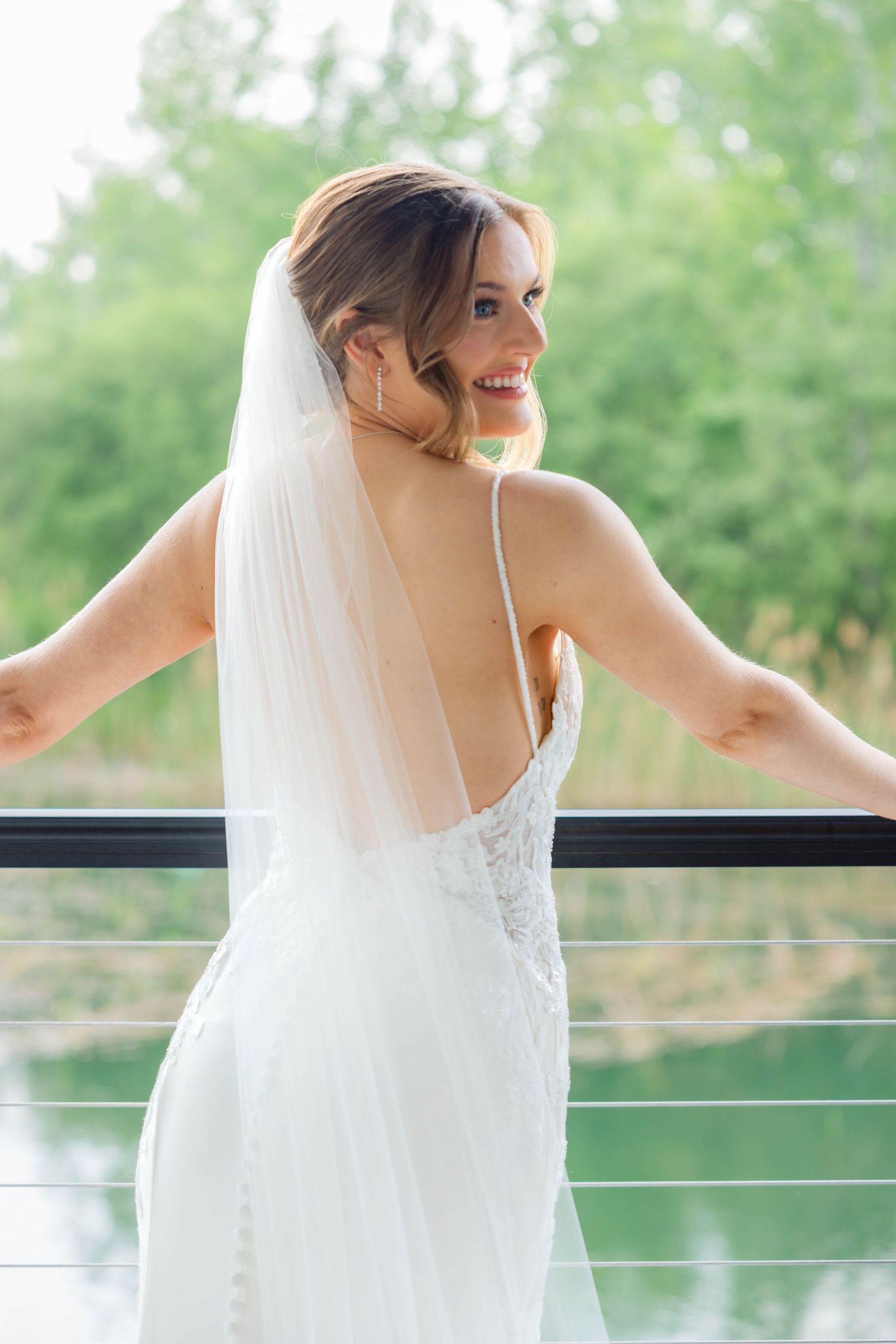 Bride in elegant backless white wedding dress with veil laughs on balcony overlooking pond.