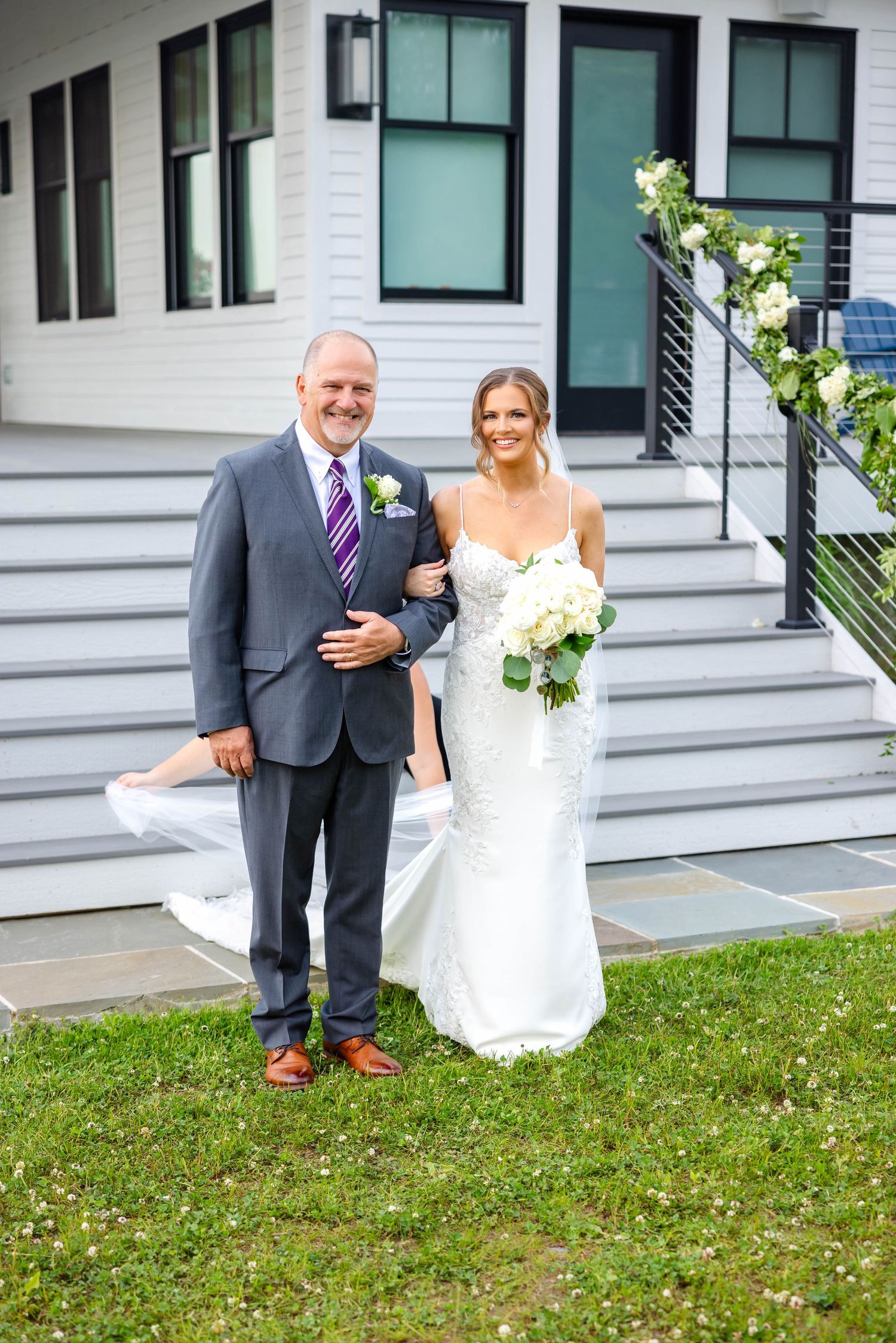 Bride in fitted lace gown holding white bouquet poses with father before outdoor wedding ceremony.