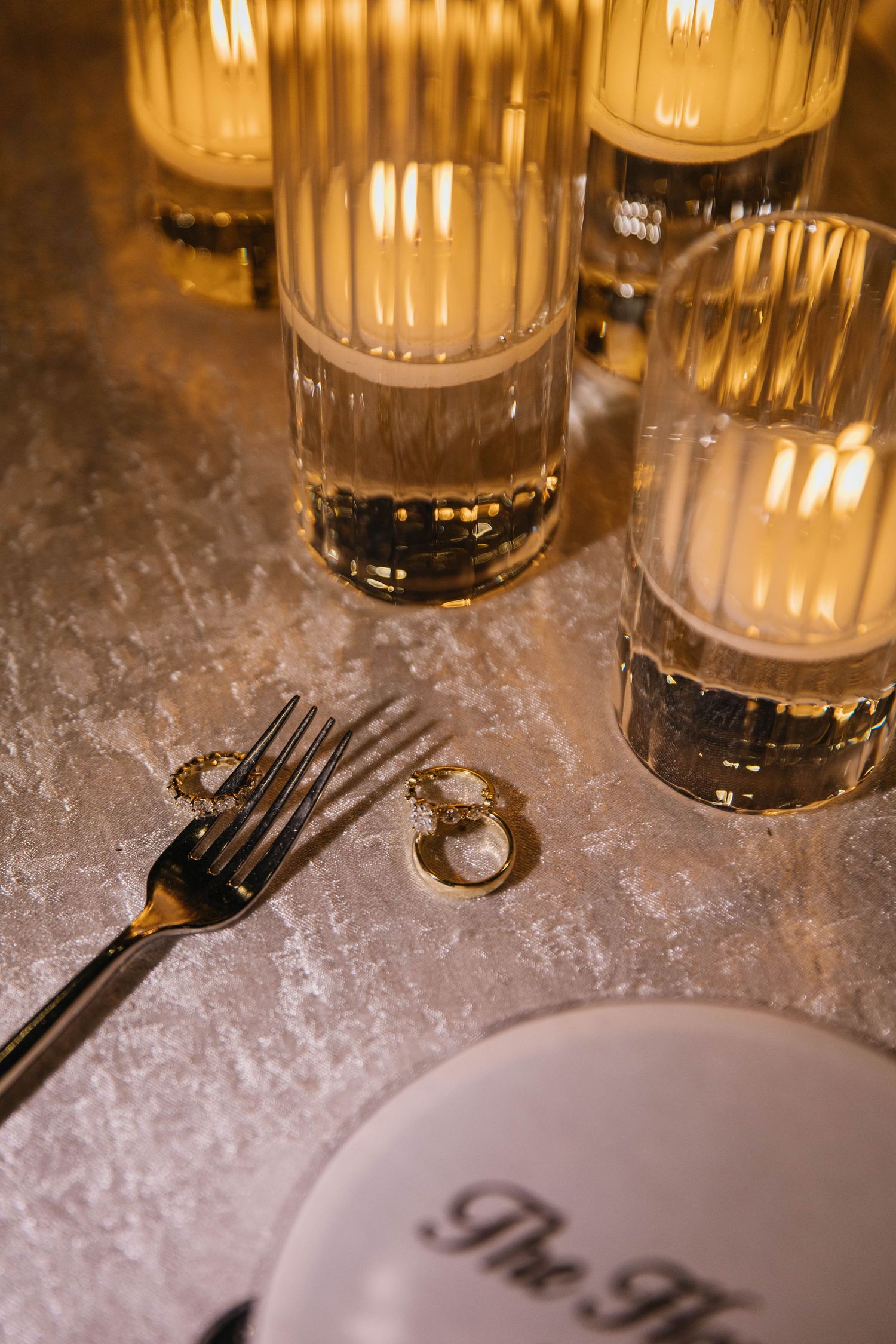 Elegant candlelit table setting with gold fork, rings, and glowing ribbed glass candle holders on a silver surface.