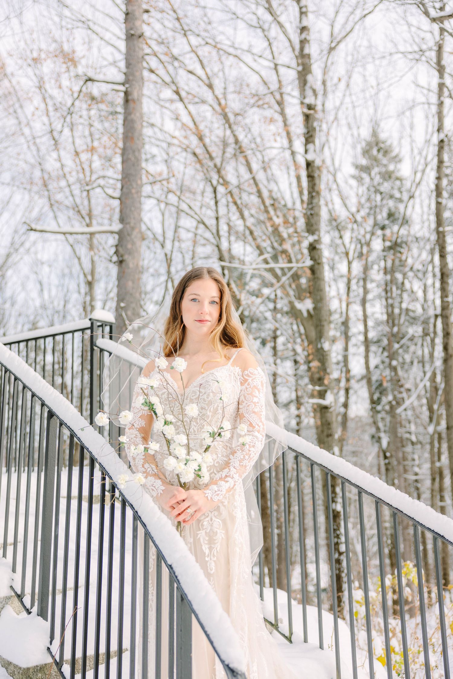 Bride in lace long-sleeve gown holding white bouquet stands on snow-covered staircase surrounded by winter trees.