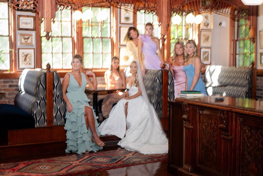 Bride in white wedding gown poses with bridesmaids in colorful dresses inside a rustic venue.