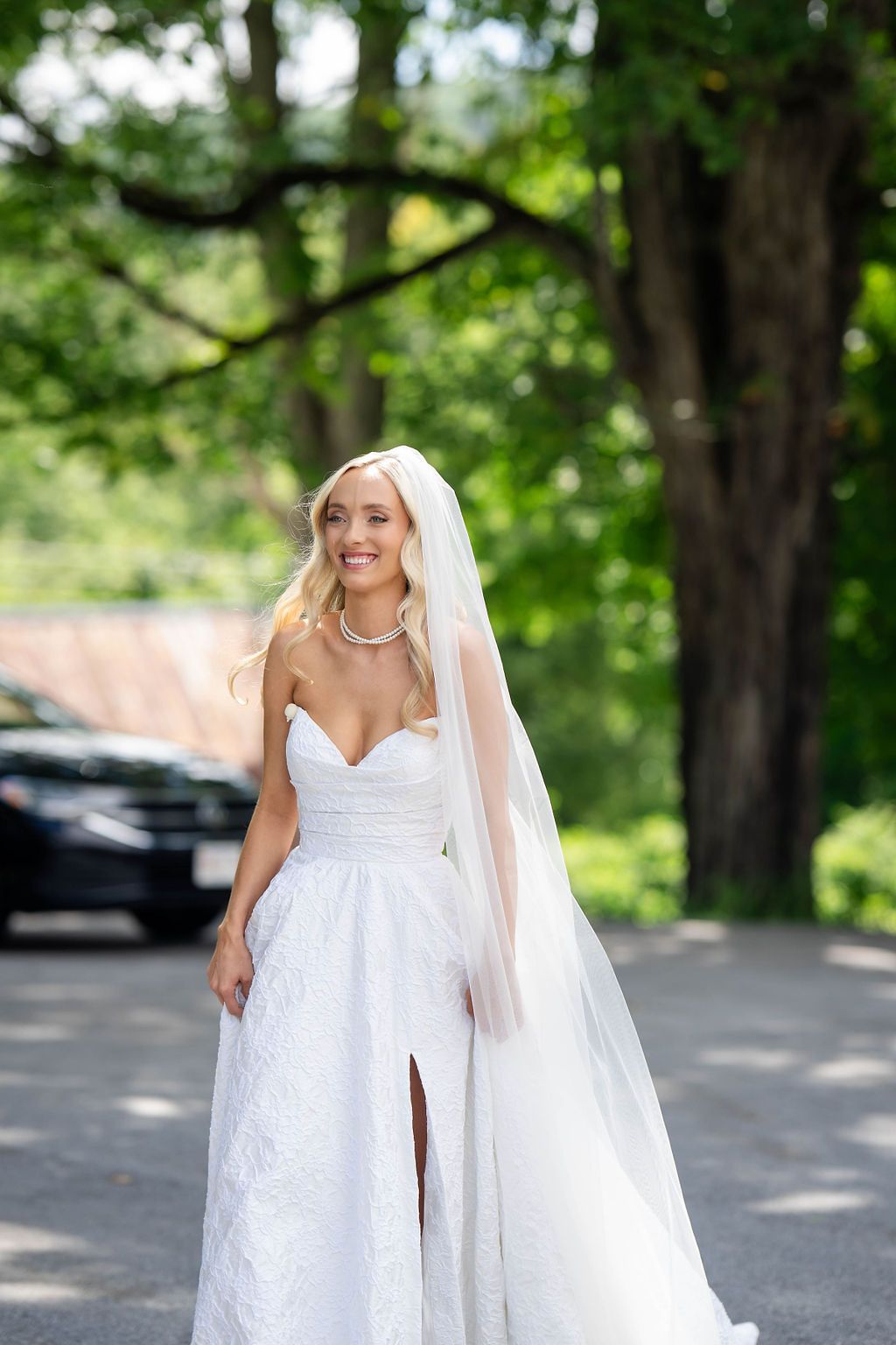 Smiling blonde bride in strapless white lace gown and veil walks outdoors among green trees on her wedding day.