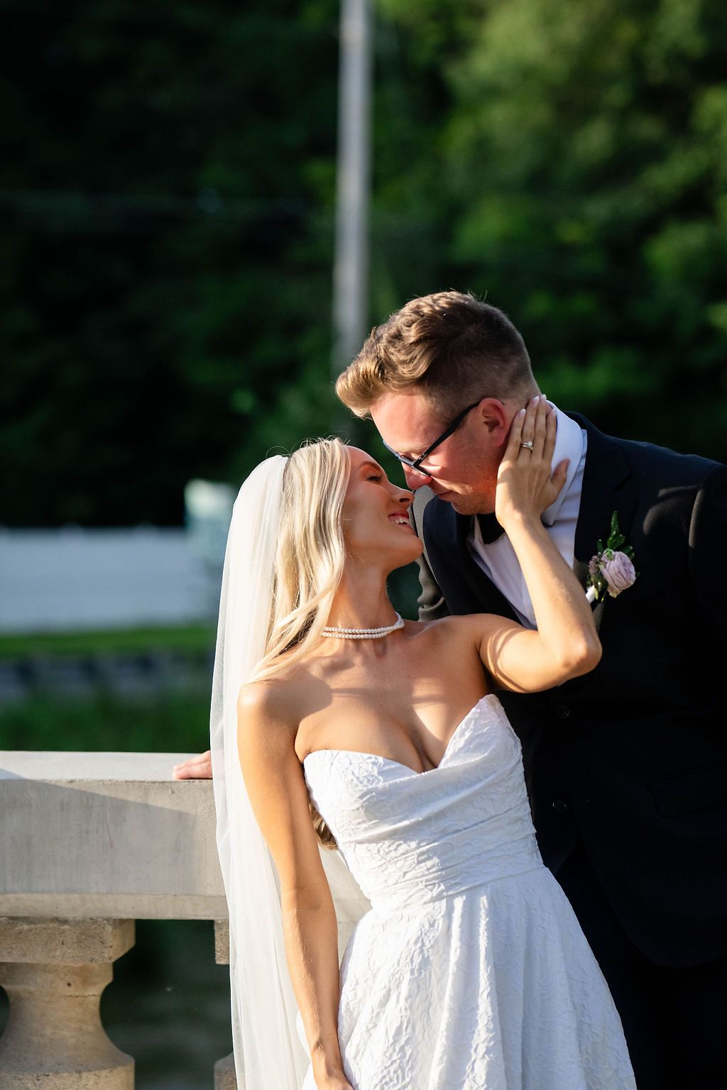 Bride in white strapless gown and veil shares a romantic kiss with groom in navy suit outdoors.