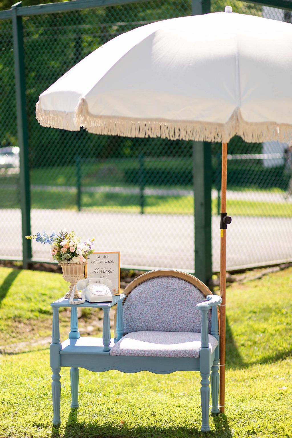Charming outdoor wedding guest book station with blue chair, white umbrella, flowers, and vintage phone.