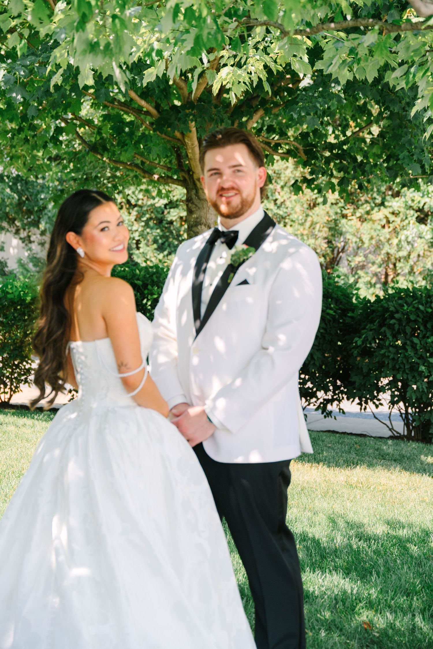 Happy bride and groom posing together under green tree in dappled sunlight at summer wedding.
