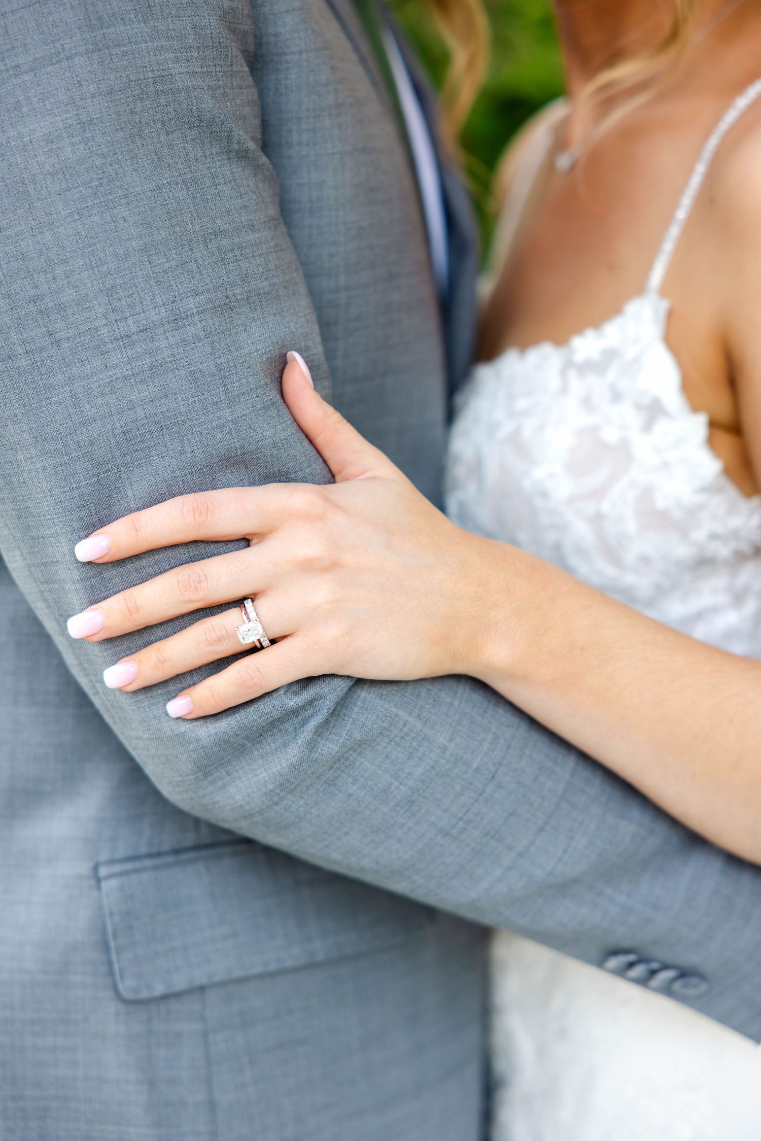 Close-up of bride's diamond wedding ring as she holds groom's arm in gray suit.