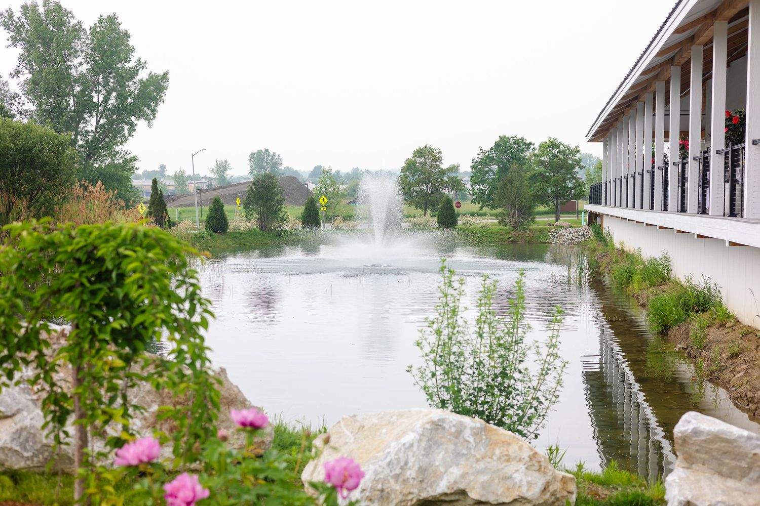 Serene wedding venue pond with a fountain surrounded by lush trees and pink flowers on a cloudy day.