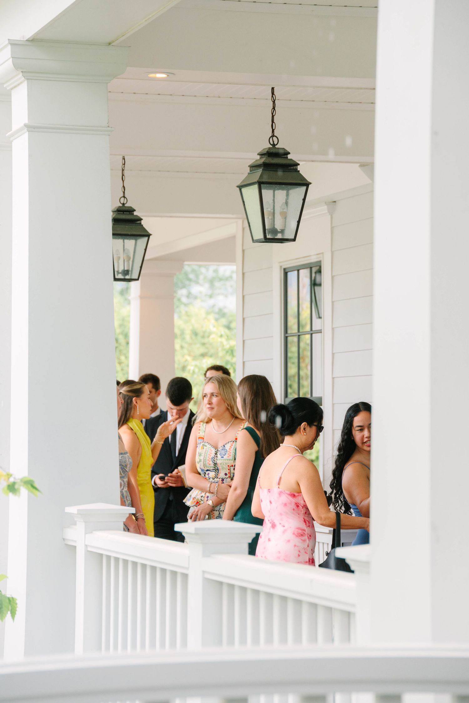 Wedding guests mingling on a classic white columned porch with hanging lanterns.
