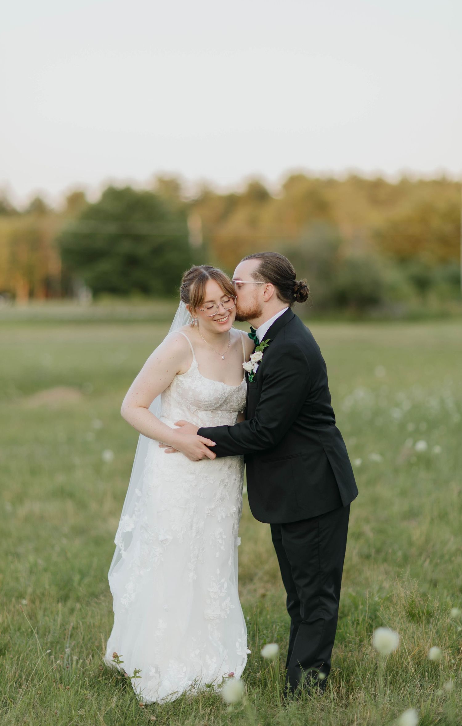 Bride and groom share a romantic kiss in a sunlit meadow, with the groom in a black suit and bride in a lace wedding gown.