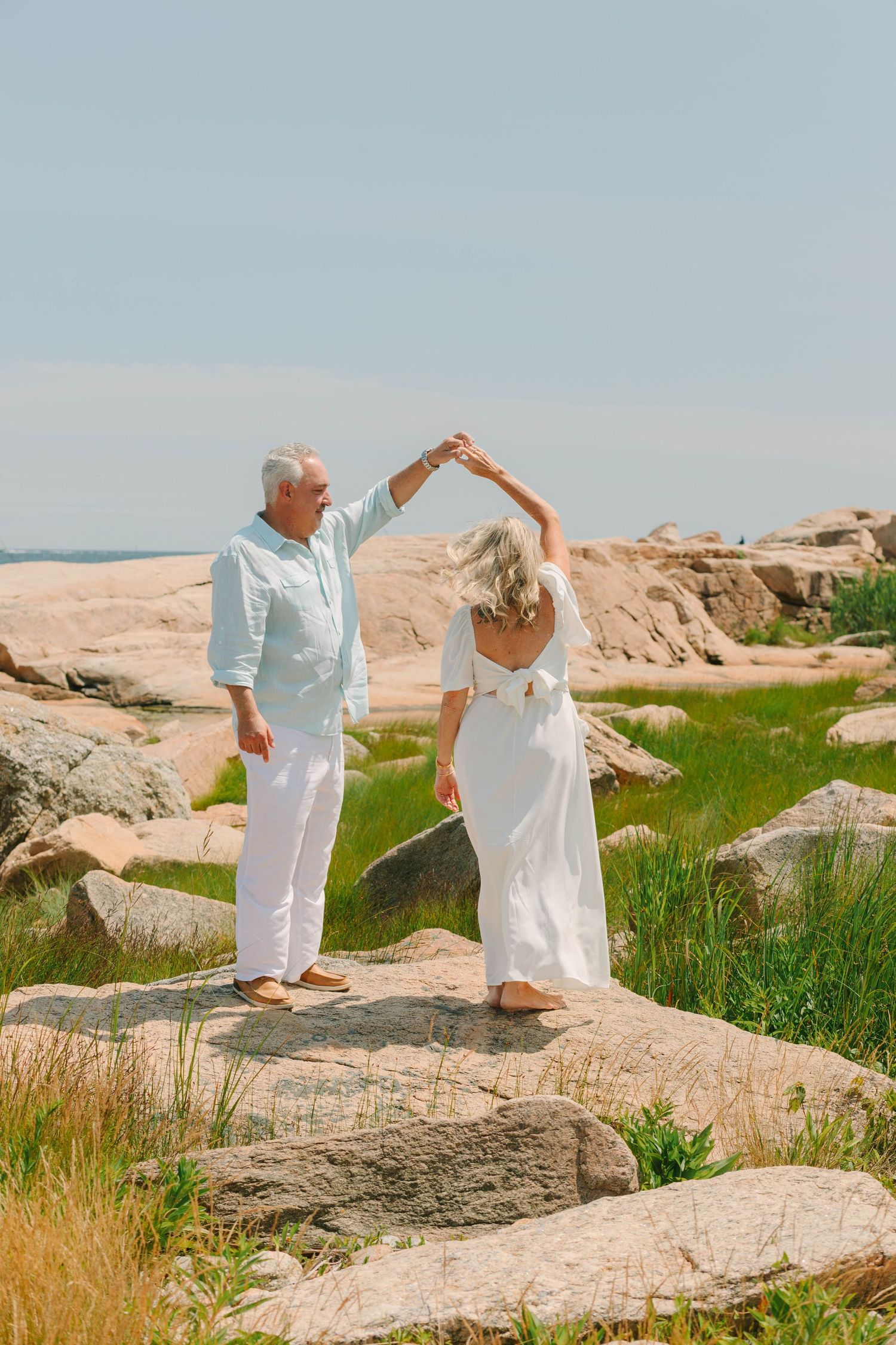 Elderly couple dancing on coastal rocks, man in blue shirt twirling woman in white dress outdoors.