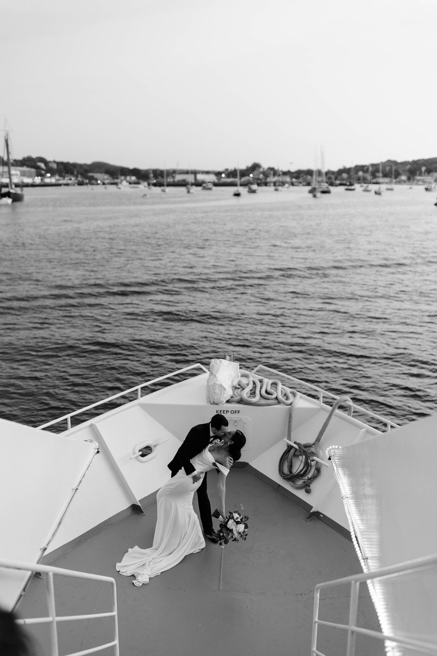 Black and white photo of bride and groom sharing romantic kiss on bow of boat with harbor backdrop.