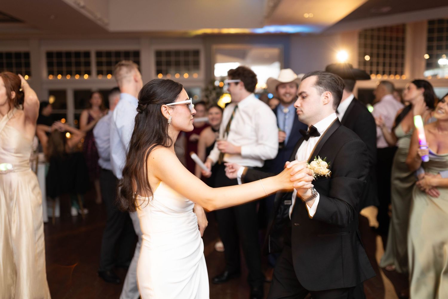 Bride in white gown and groom in tuxedo dance together wearing fun glasses at wedding reception.