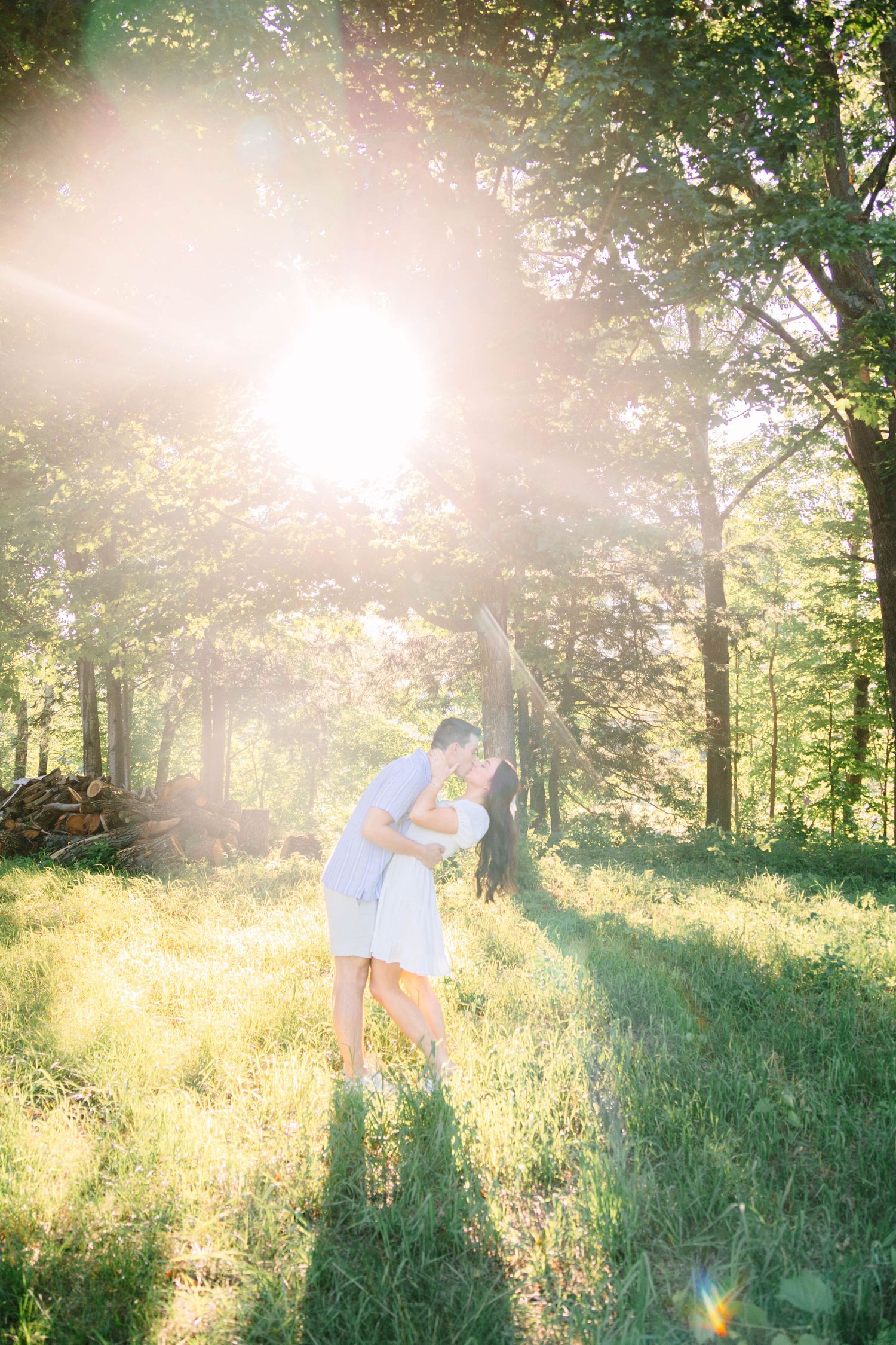Couple sharing a romantic kiss in a sunlit forest clearing, surrounded by lush green trees and glowing golden light.