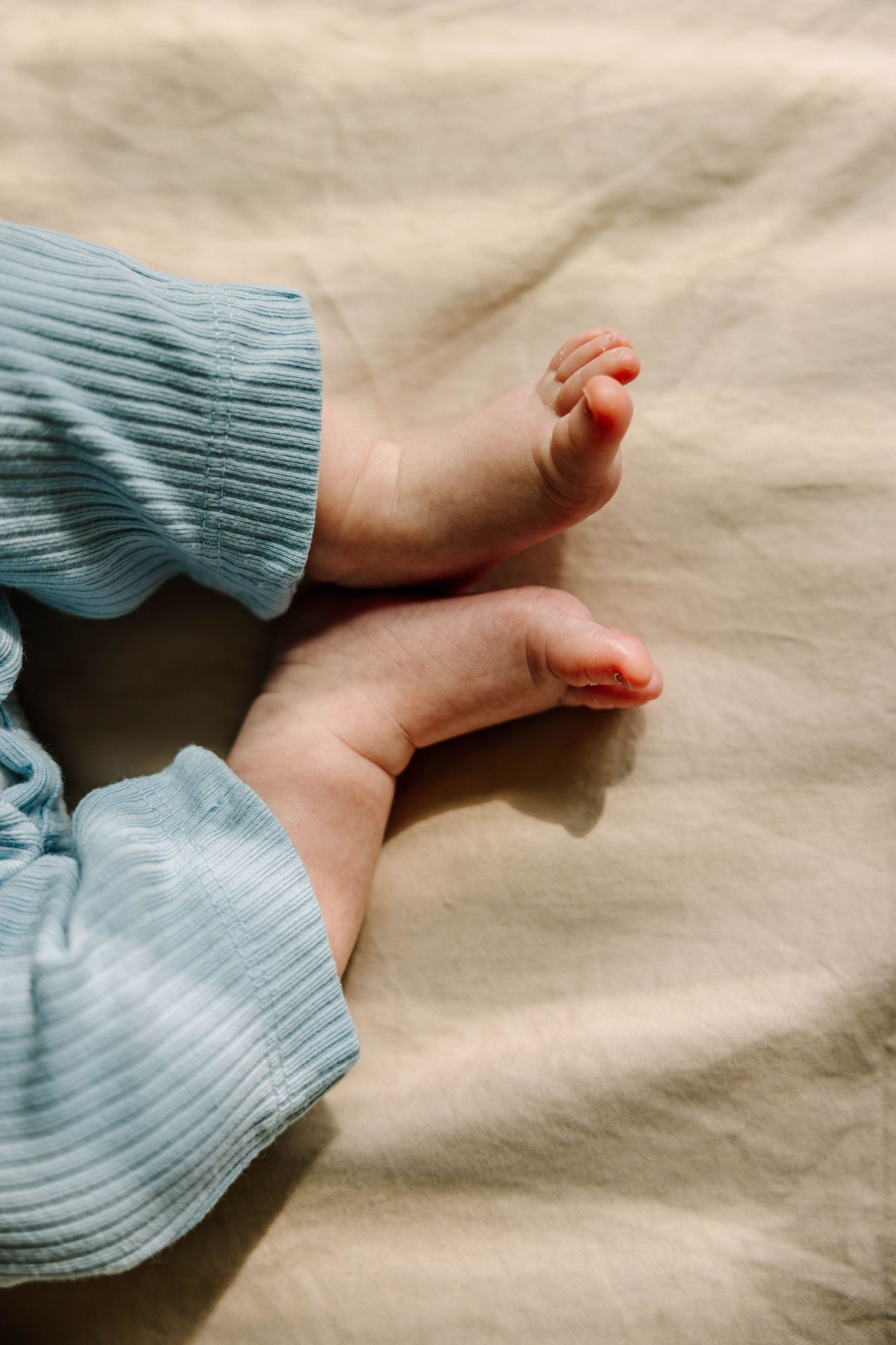 Close-up of tiny newborn baby feet wearing a light blue ribbed outfit, resting on a beige linen blanket.